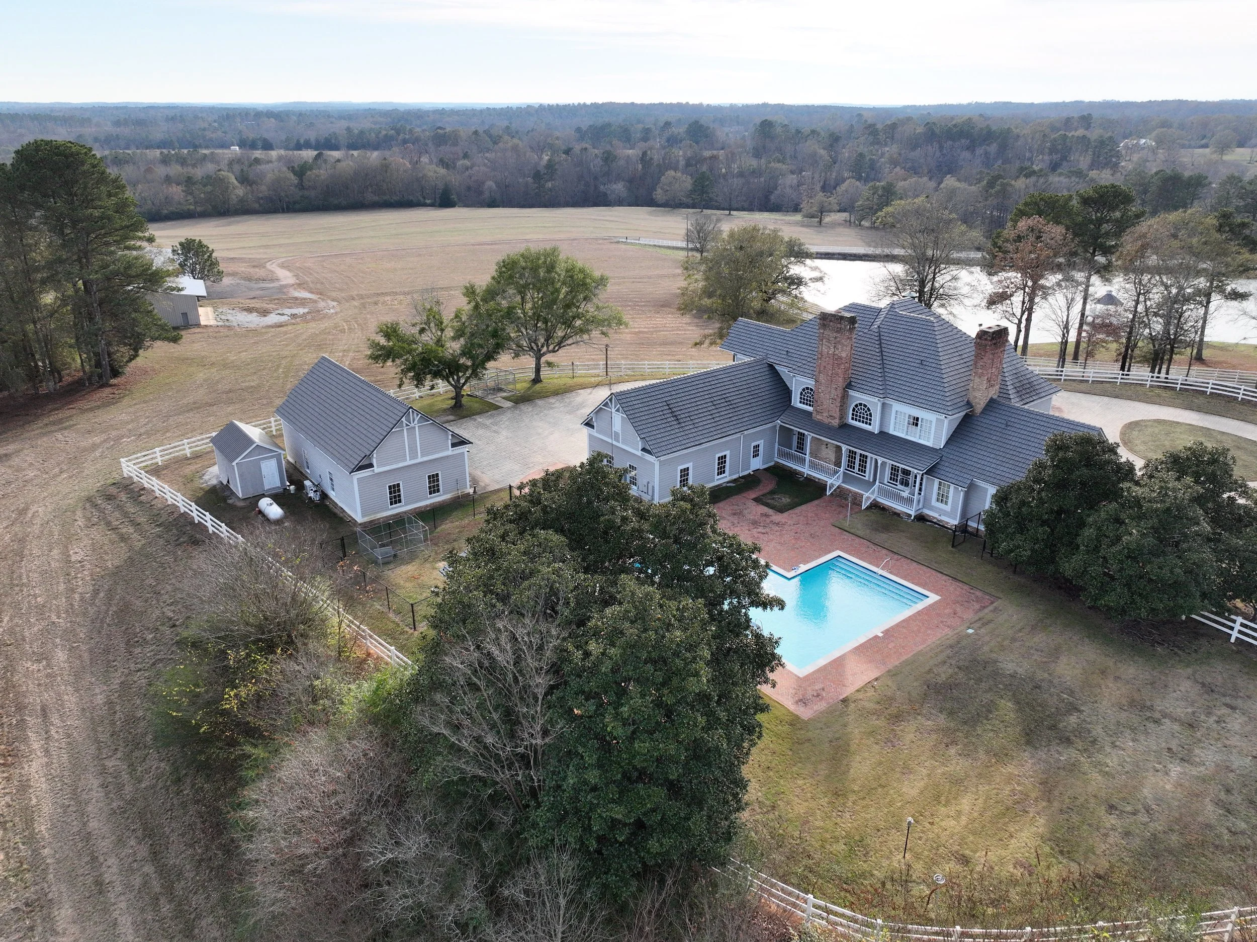 A large gray house with a swimming pool in the backyard, surrounded by trees and fencing, located on a spacious property with open fields and a pond in the background.