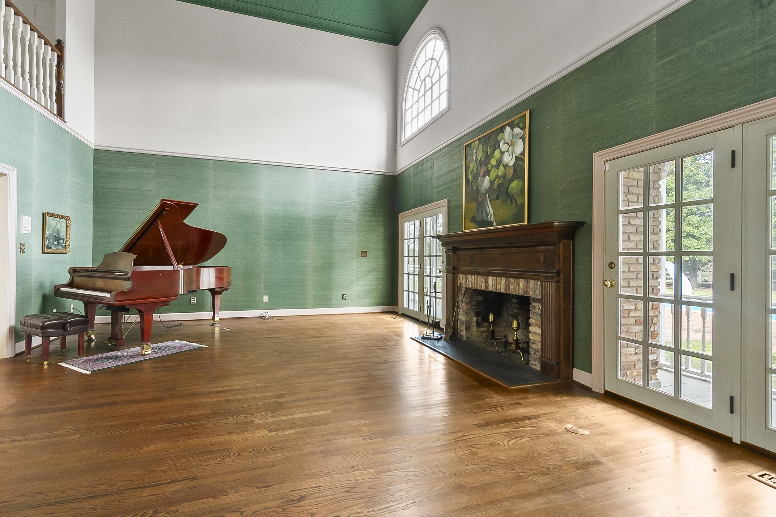 Living room with hardwood floors, green textured walls, a brick fireplace with wood mantel, a painting above the mantel, glass-paneled double doors, and a grand piano.