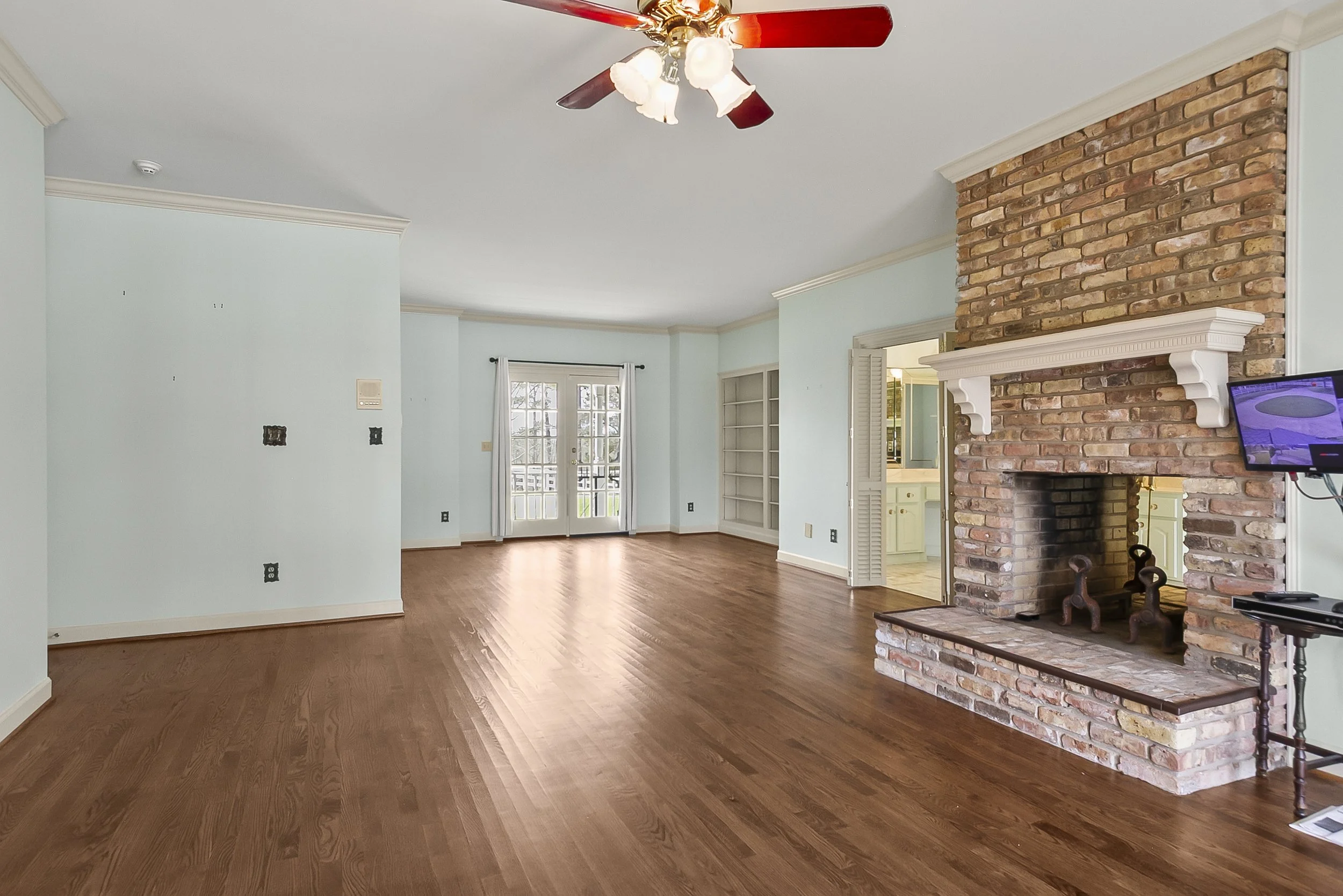 Empty living room with hardwood floors, a brick fireplace, a ceiling fan with lights, and a set of French doors leading outside.