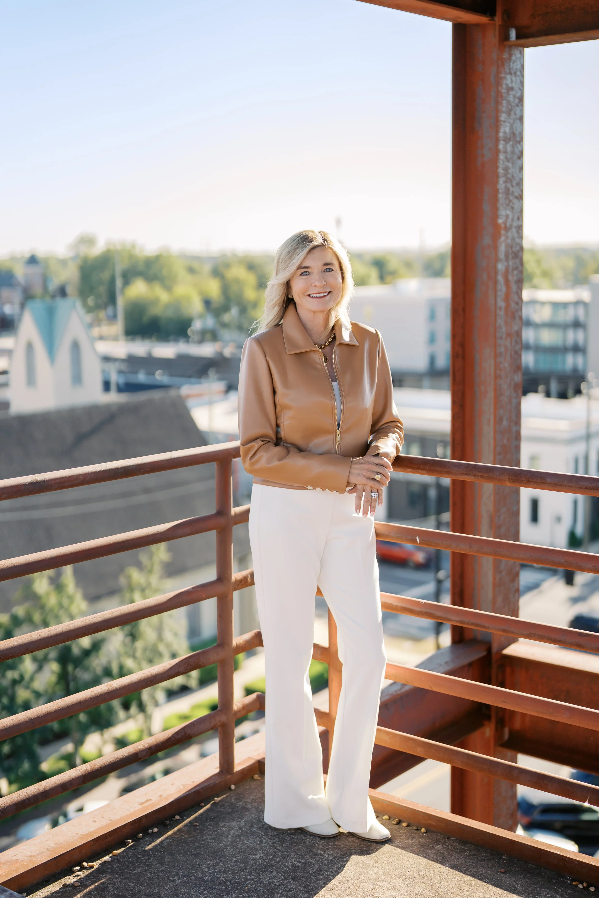A smiling woman with blonde hair standing on a rooftop balcony, wearing a tan leather jacket and white pants, with a cityscape in the background during daylight.