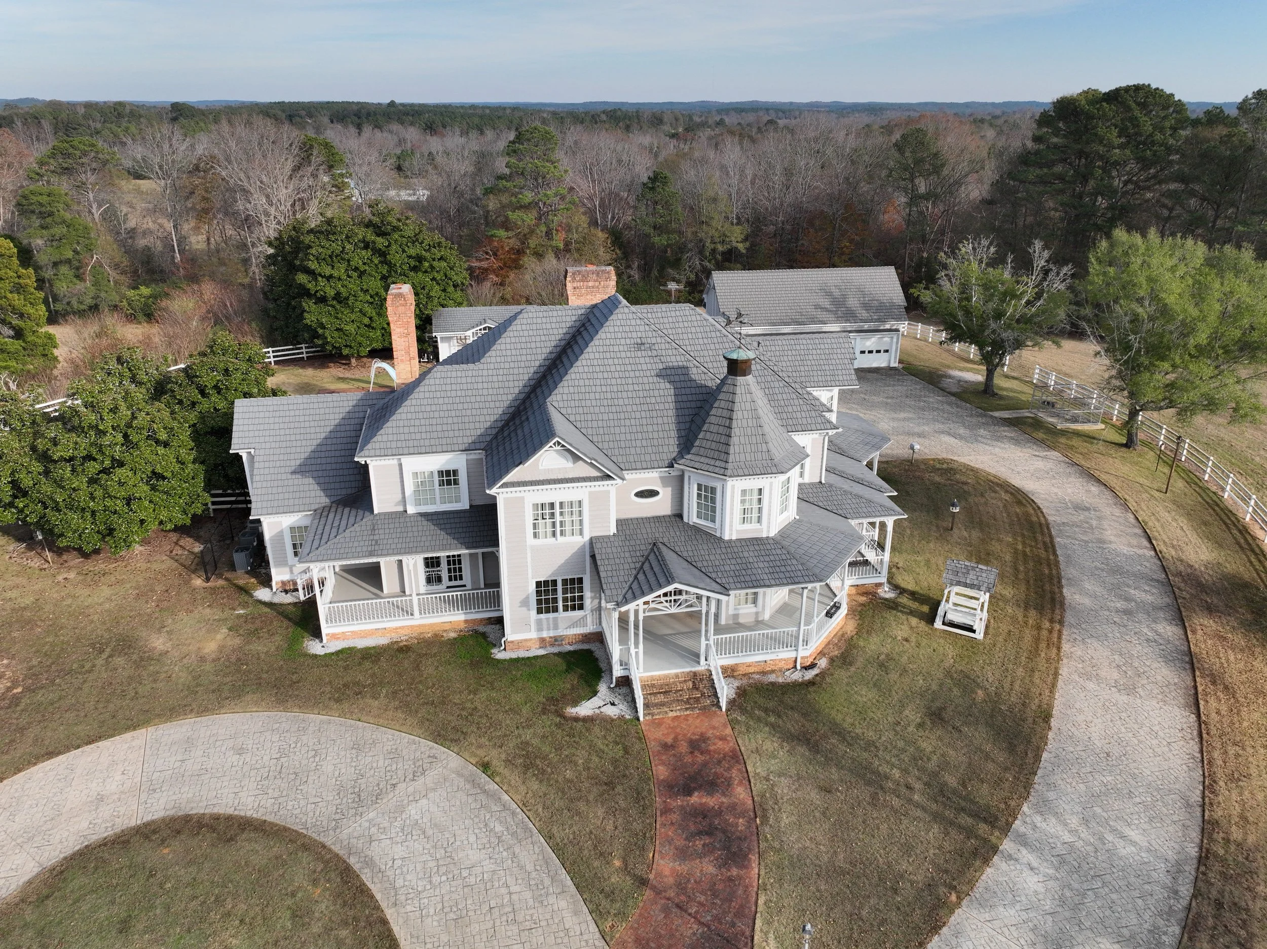 A large, white, multi-story house with a gray roof, surrounded by trees and a curved driveway, located in a rural area with wooded land in the background.