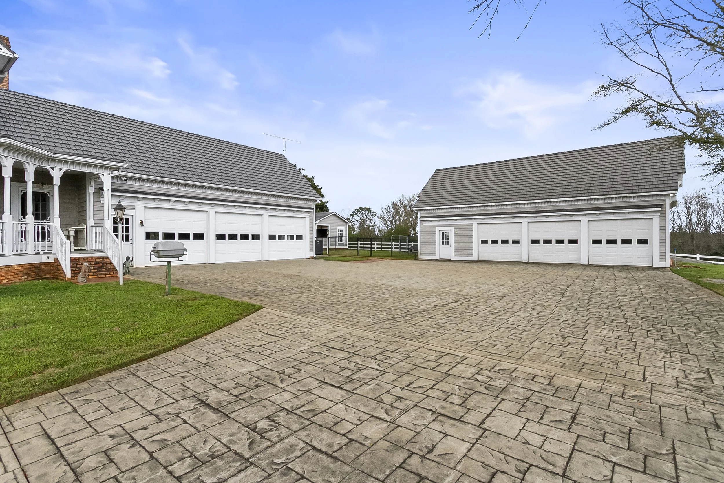 A spacious driveway with brick paving leading to two white garage buildings with multiple doors, a small grassy area with a mailbox, and a white fence in the background under a clear blue sky.
