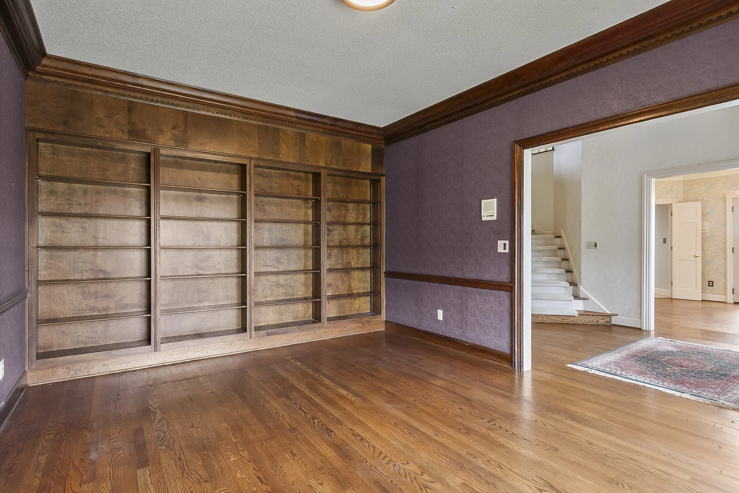 Empty living room with hardwood floors, purple and white walls, built-in wooden shelves, and an open doorway showing a staircase.