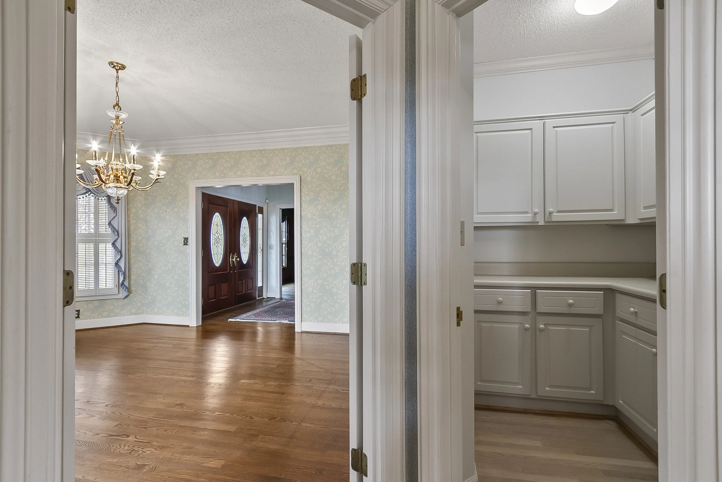 View from a doorway into a kitchen on the right with white cabinets, into a living room with wooden floors, a chandelier, and a fireplace with double front doors in the background.