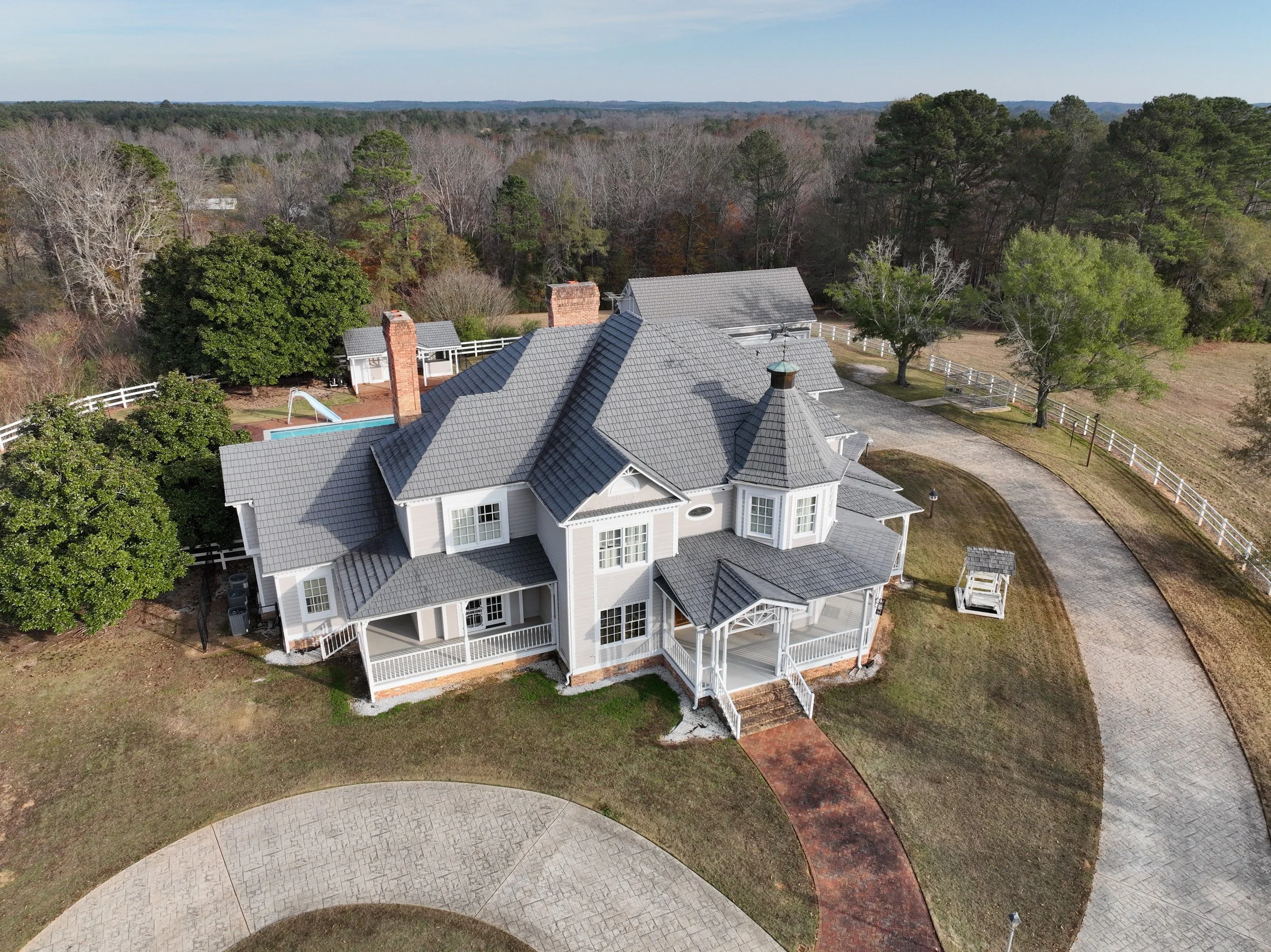 Aerial view of a large, white Victorian-style house with a gray roof, surrounded by a curved cobblestone driveway, green lawn, trees, and a white fence, with a backyard pool and a small shed.