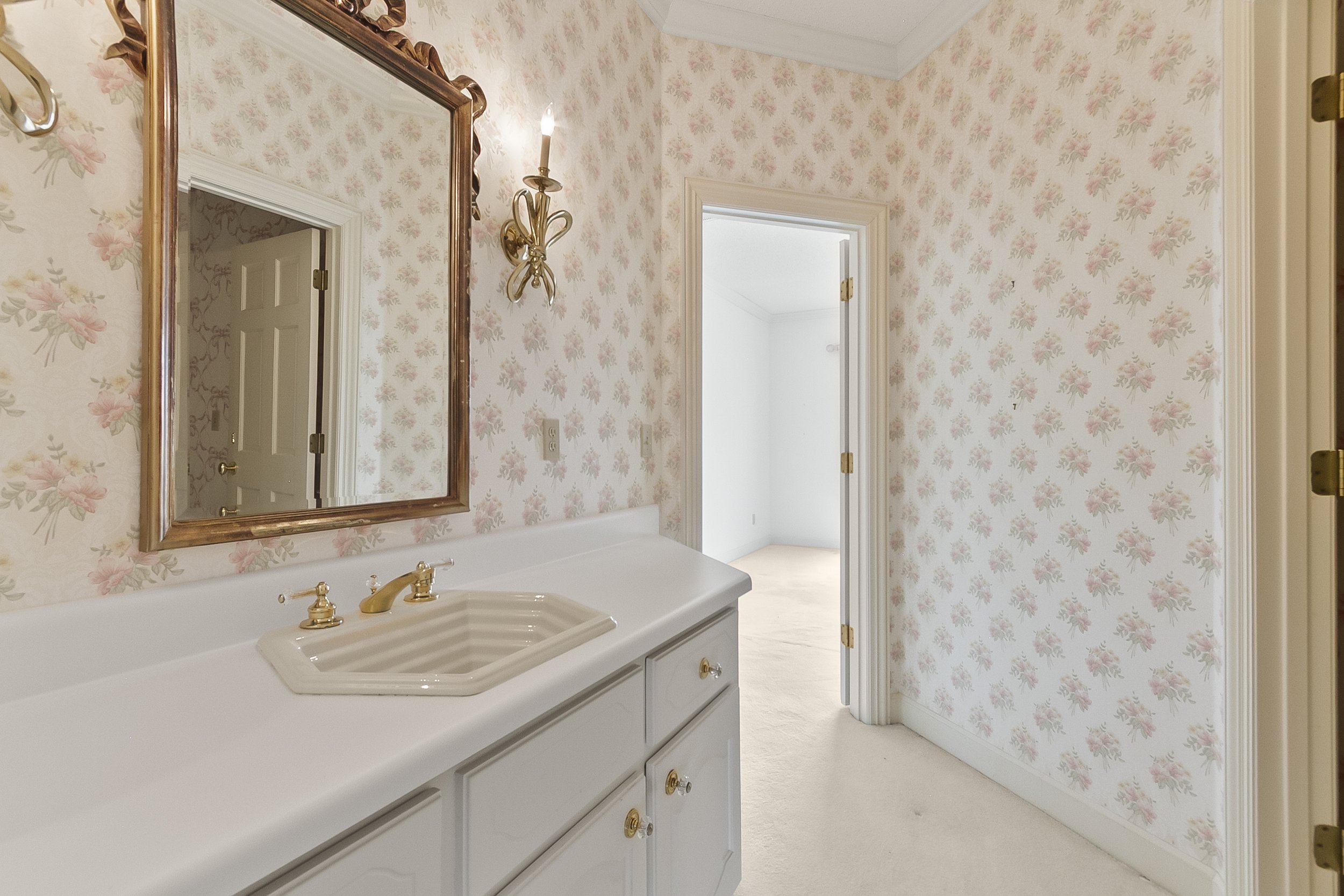 A vintage bathroom with floral wallpaper, a white vanity with a built-in sink, gold fixtures, a large mirror with a decorative wooden frame, a brass wall sconce, and an open door leading to a bright room.