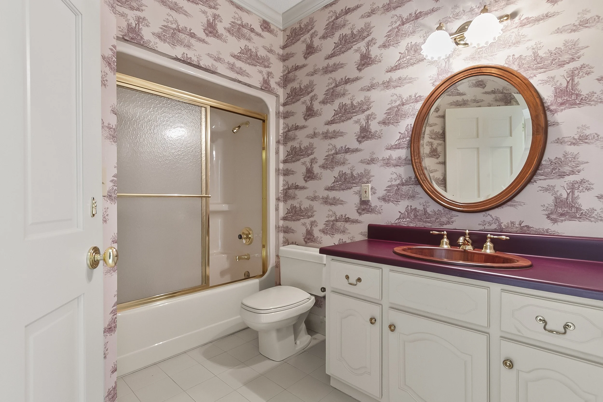 Bathroom with floral pink wallpaper, white cabinet with burgundy countertop, round mirror, gold fixtures, toilet, and a bathtub with a frosted sliding door.