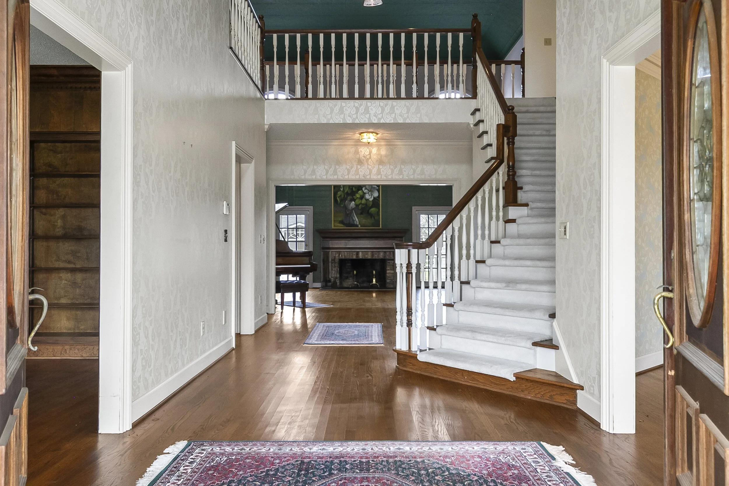 Entryway with wooden door open, hardwood floor, staircase with white carpet and wooden handrail, and a living room with a fireplace in the background.