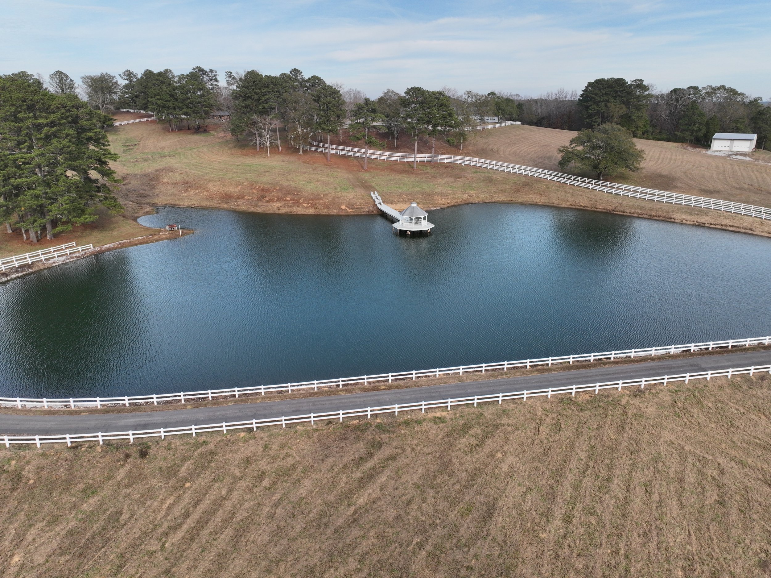 A small pond with a gazebo and dock, surrounded by a white fence, in a rural landscape with trees and fields.
