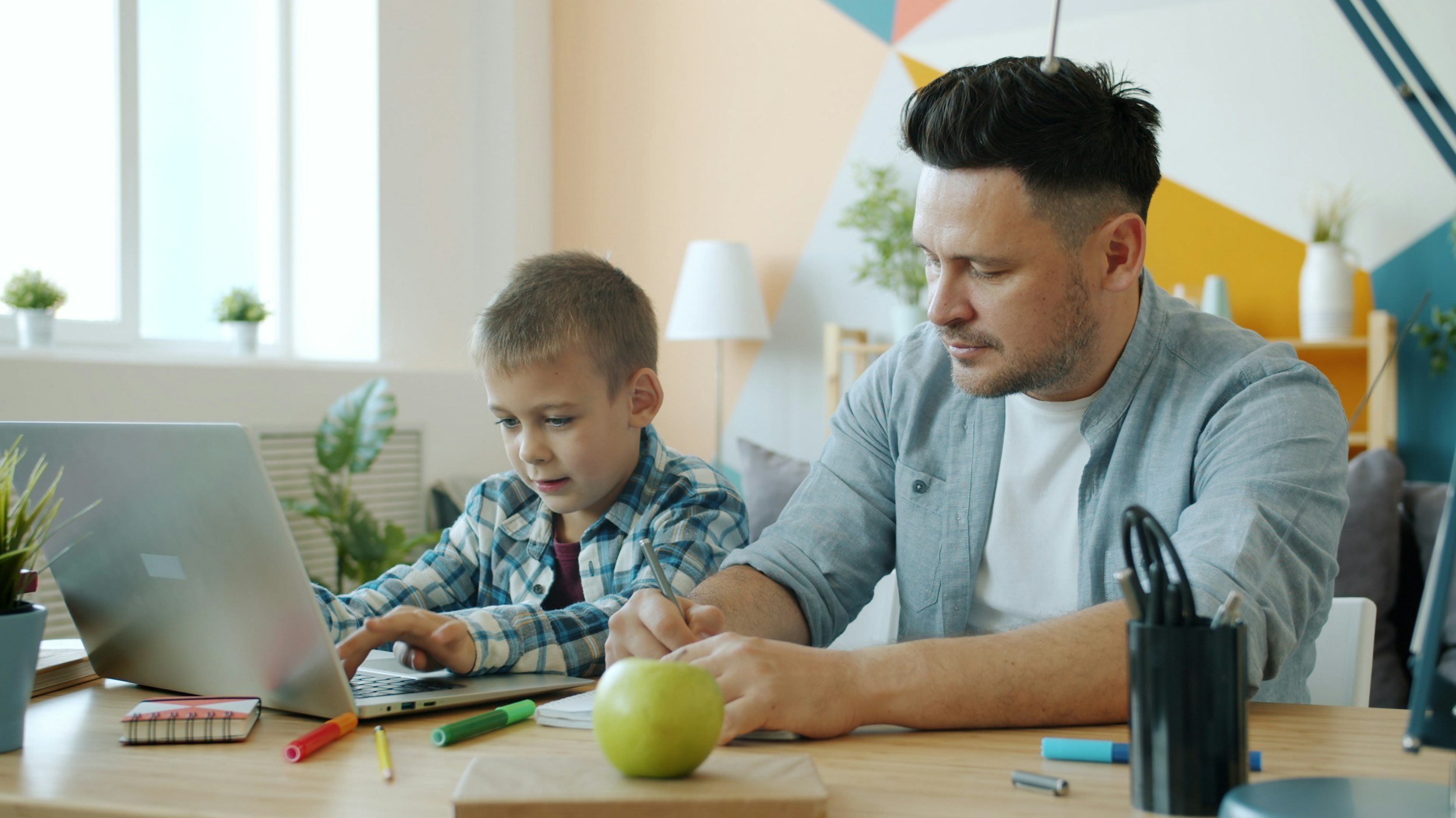 A man and a boy working on a laptop and writing at a desk with colored markers and an apple in a bright, modern room.