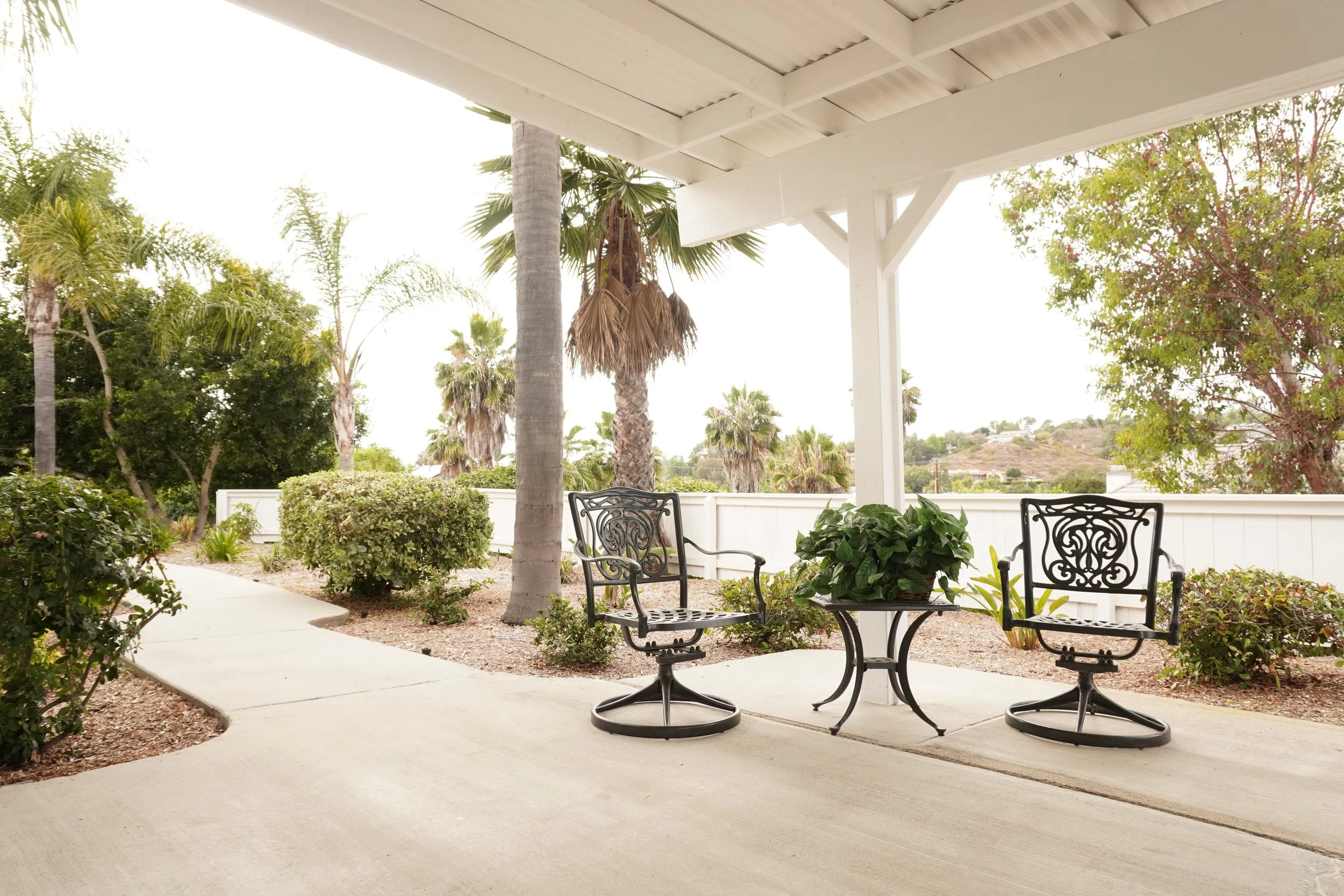 A covered patio with black metal patio furniture, including a table and two chairs, surrounded by plants, trees, and shrubs, with a white fence and a hillside in the background.
