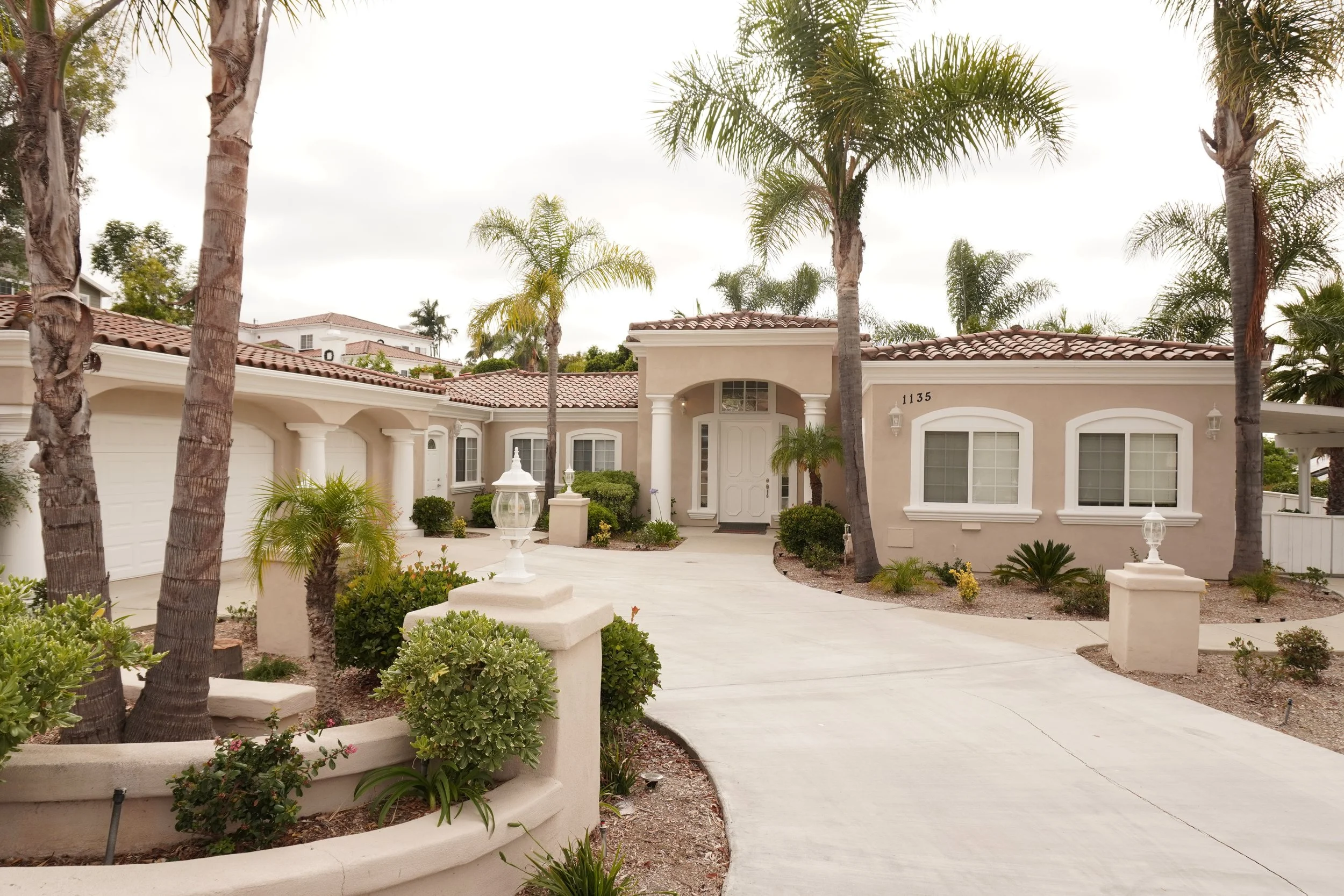 A house with a beige exterior, terracotta roof tiles, and a white front door is surrounded by palm trees and landscaped plants. There is a driveway and decorative lamps near the entrance.