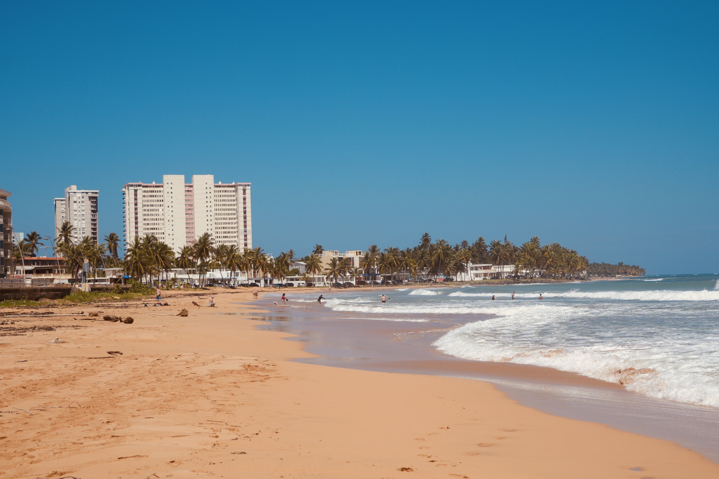 A sandy beach with people swimming and walking by the shoreline, tall buildings in the background, and clear blue sky above.