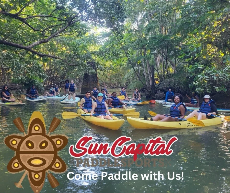 A group of people kayaking on a river surrounded by dense green trees, with some people smiling at the camera. There is a logo with a paddle and turtle, and text that reads 'Sun Capital Paddlesports Come Paddle with Us!'