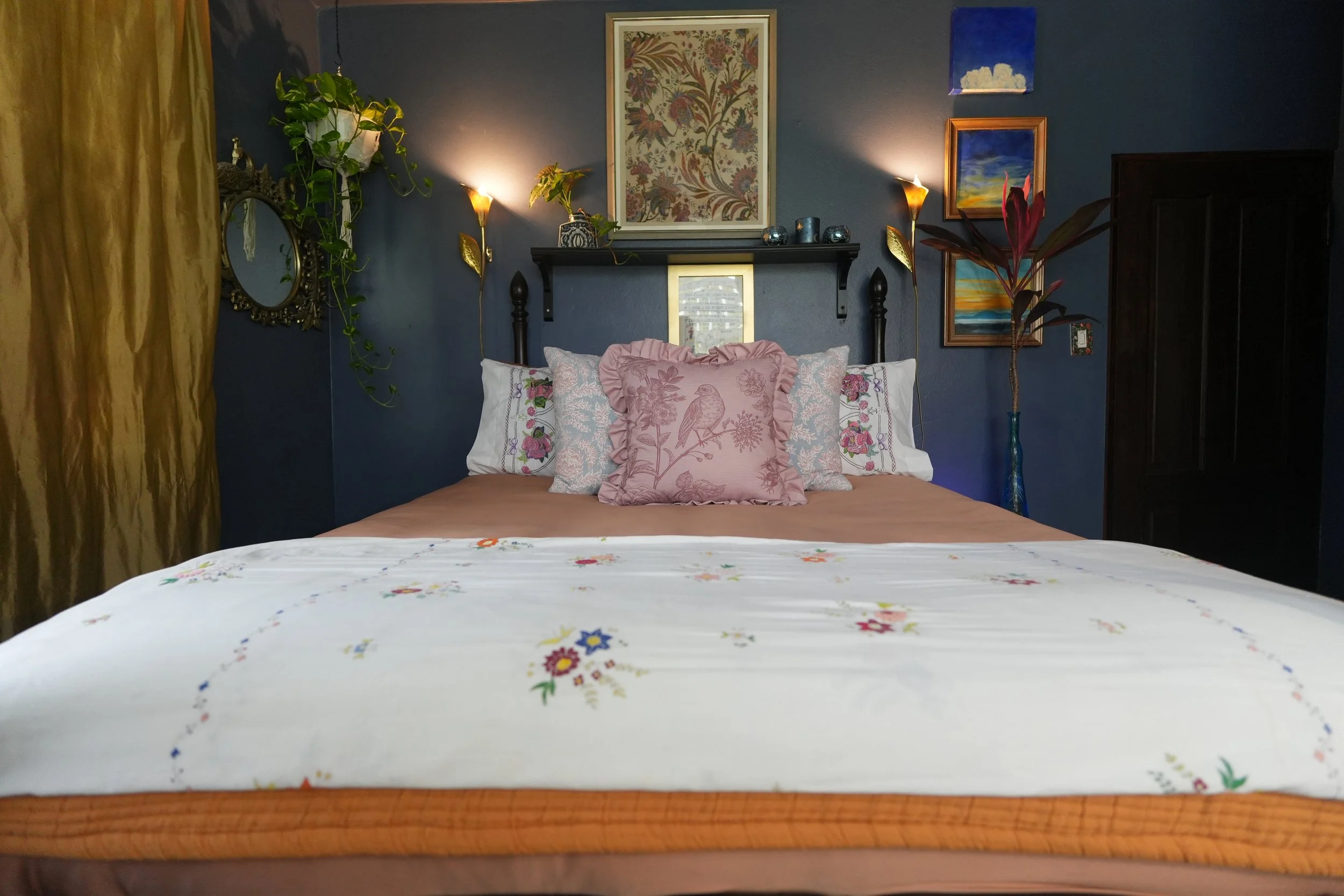 A bedroom with navy blue walls, a bed with floral embroidered bedding and pillows, a wooden headboard, framed artworks on the wall, hanging plants, lamps, and gold curtains on the left side.