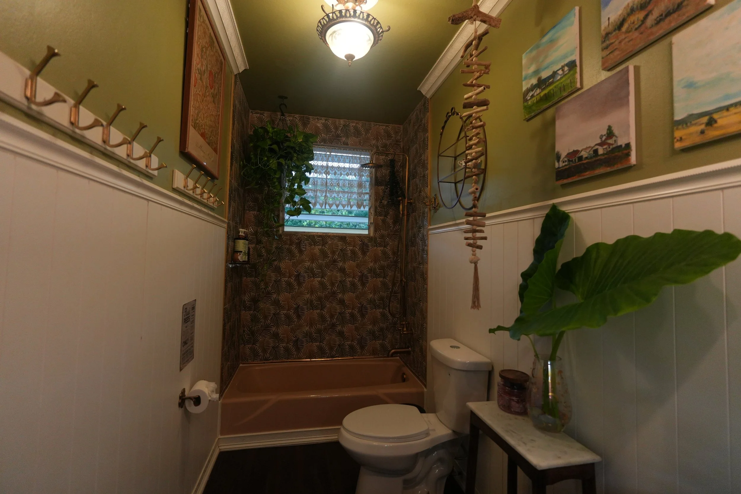 A small bathroom with green walls and white wainscoting. There is a window with patterned curtains, a potted plant, and framed artwork on the walls. A floating shelf with decorative items and a hanging driftwood art piece are visible. The bathroom ha