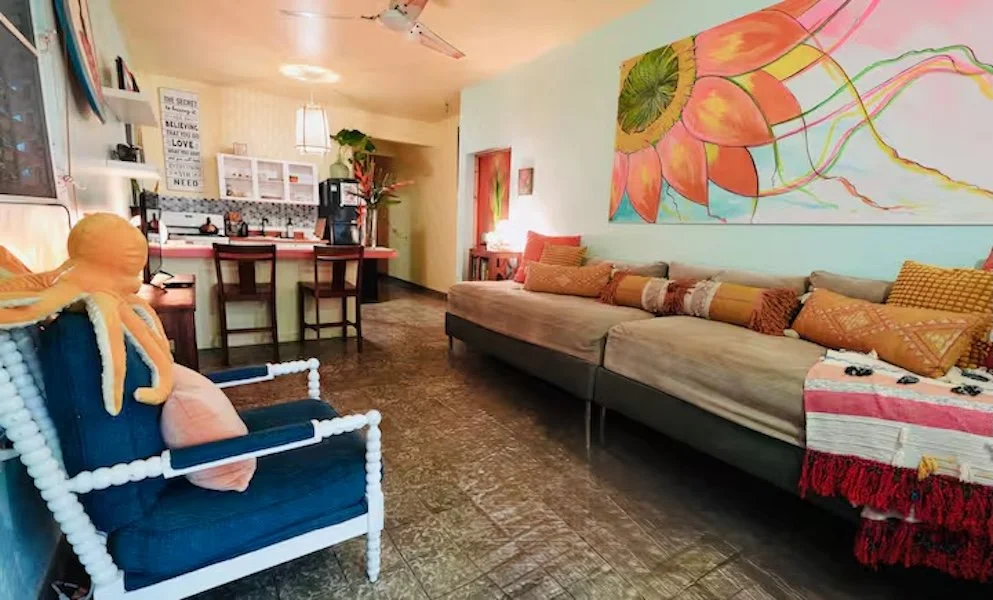 Living room with a blue armchair, mustard-colored pillows, an L-shaped sofa with various pillows, colorful wall art, and a small bar area with two barstools.