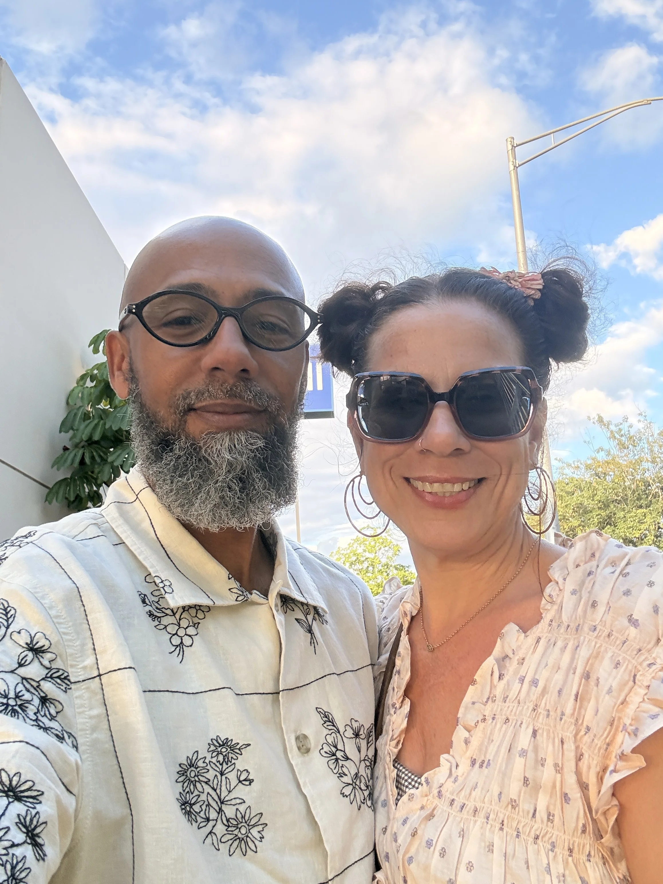 A smiling man and woman taking a close-up selfie outdoors on a sunny day with a blue sky and clouds in the background.