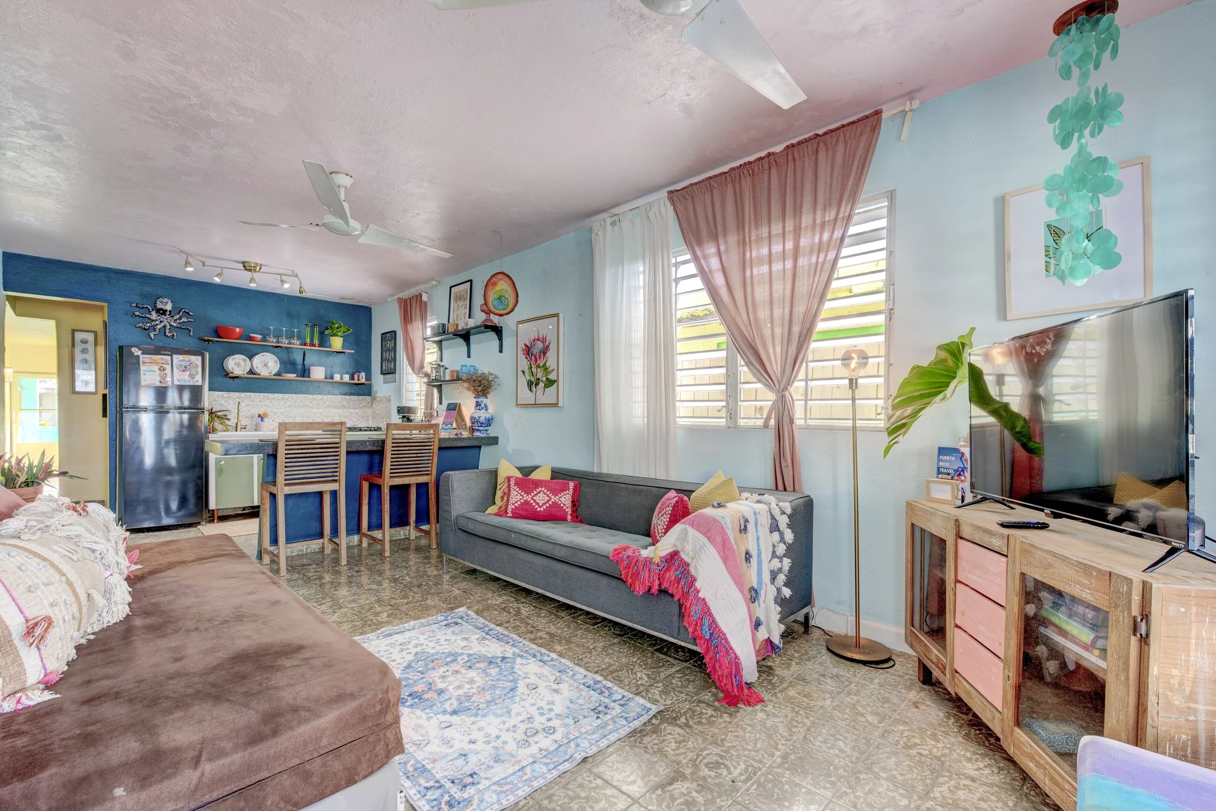 A cozy living room with pastel blue walls, large windows with white and pink curtains, and a TV on a wooden stand. The room has a gray sofa with red and yellow decorative pillows, and a brown throw blanket. There are decorative items on the walls, including a framed picture and a wall art with a rainbow. The ceiling has white fans and track lighting. A colorful area rug covers part of the floor, and there are various plants and decorative items, such as a blue vase, hanging on the walls.