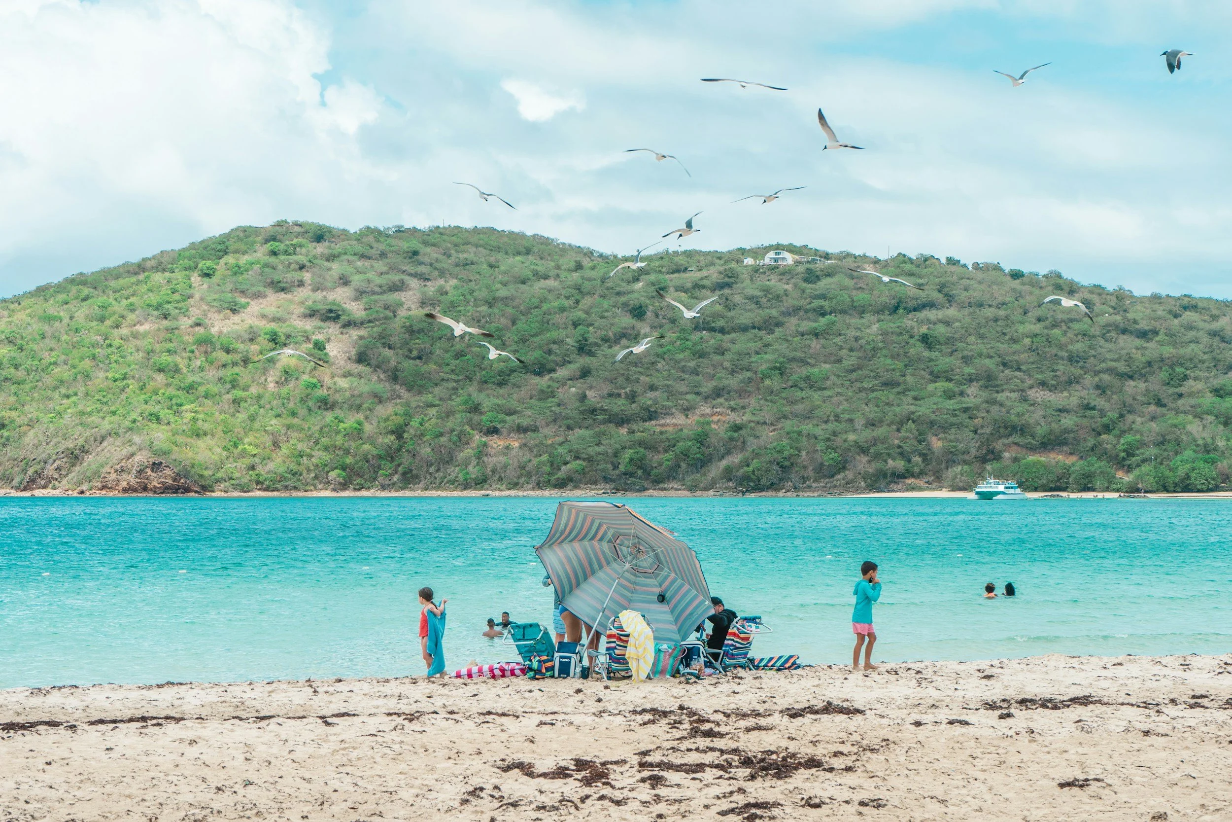 People relaxing on a sandy beach in Culebra with an umbrella, children playing near the water, seagulls flying above, a boat in the distance, and green hills with a house on top under a partly cloudy sky.