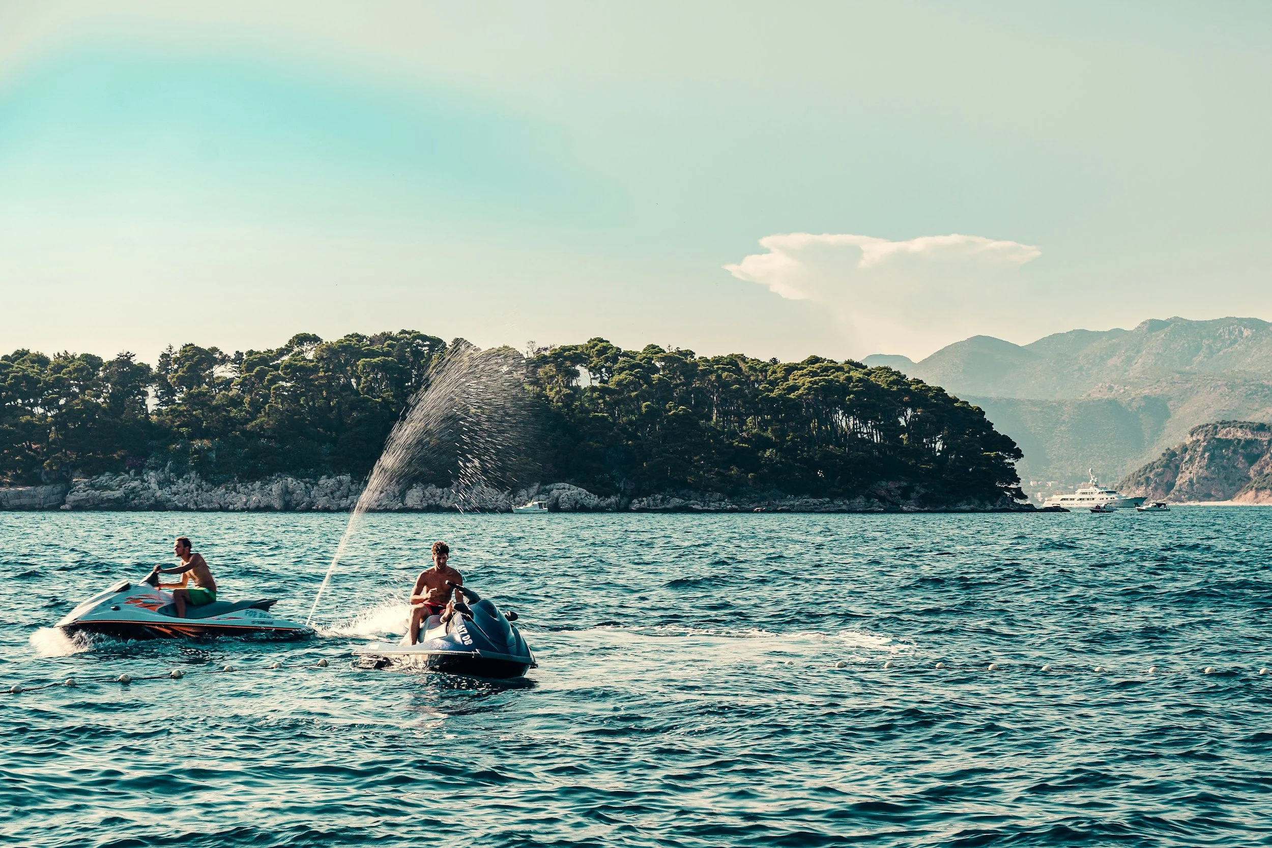 Two people riding jet skis on a body of water near a tree-covered island with mountains in the background.