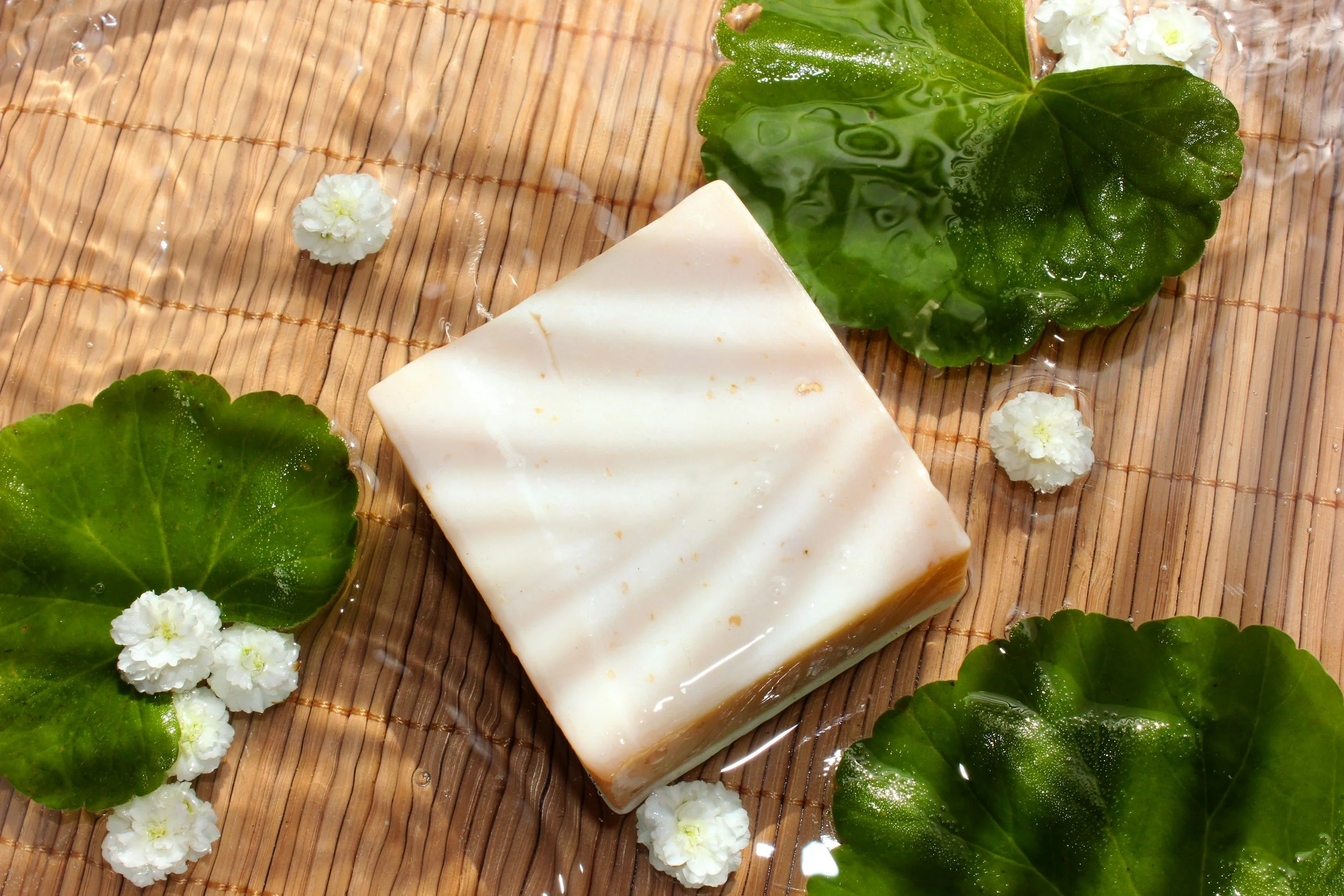 A bar of soap on a bamboo mat surrounded by large green leaves, small white flowers, and water droplets.