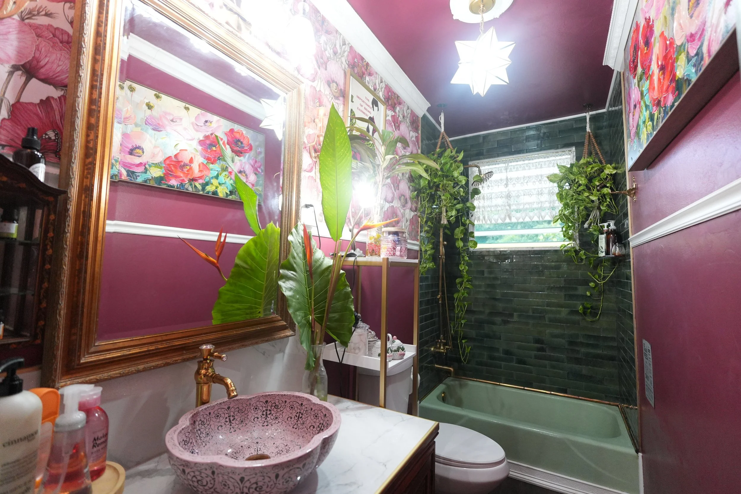 A bathroom with pink walls, decorated with floral wallpaper and artwork. There is a gold-framed mirror above a marble countertop with various bottles and a pink decorative bowl. Green plants hang near the window with patterned curtains, and a bathtub