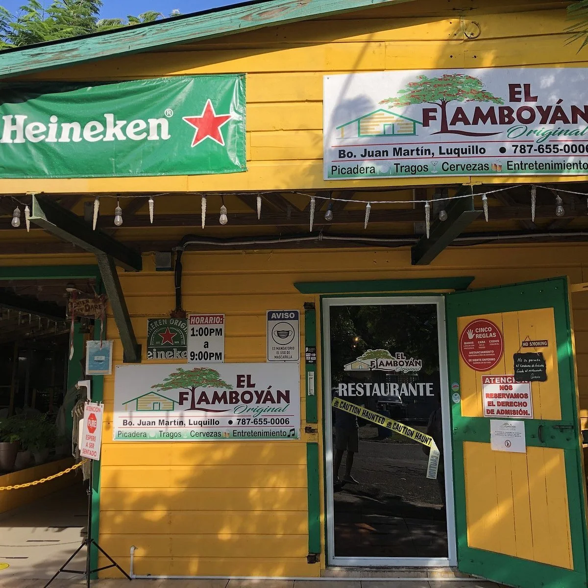 Yellow building with green accents, multiple signs including a Heineken advertisement, a sign for El Famboyán restaurant, and various notices and signs near the entrance, including operation hours, safety protocols, and caution tape.