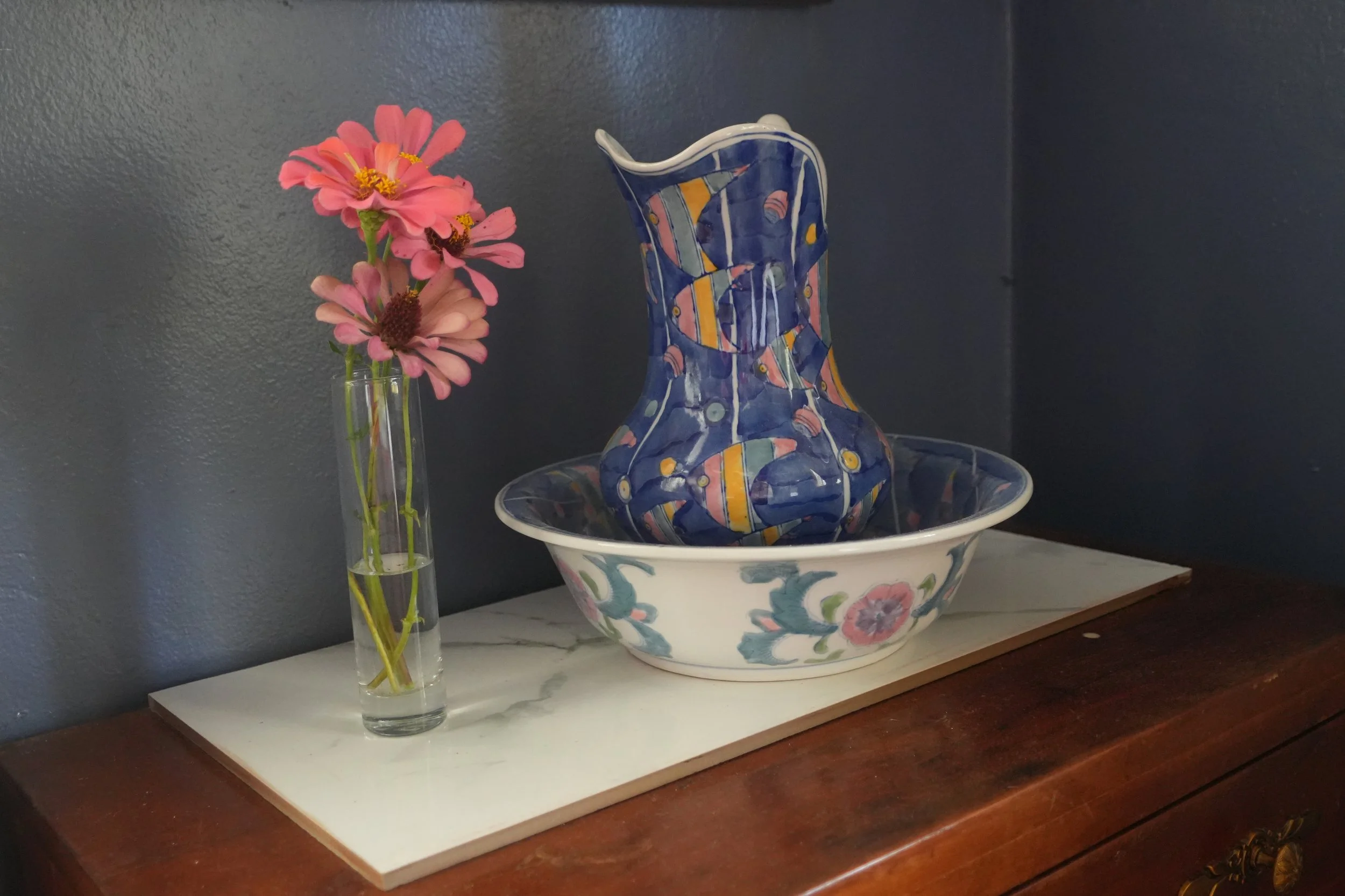 A tall, narrow glass vase with pink zinnia flowers on a white marble surface. Next to it, a decorative ceramic pitcher with an abstract, colorful pattern, sitting inside a matching ceramic bowl with floral designs. The background is a dark gray wall.