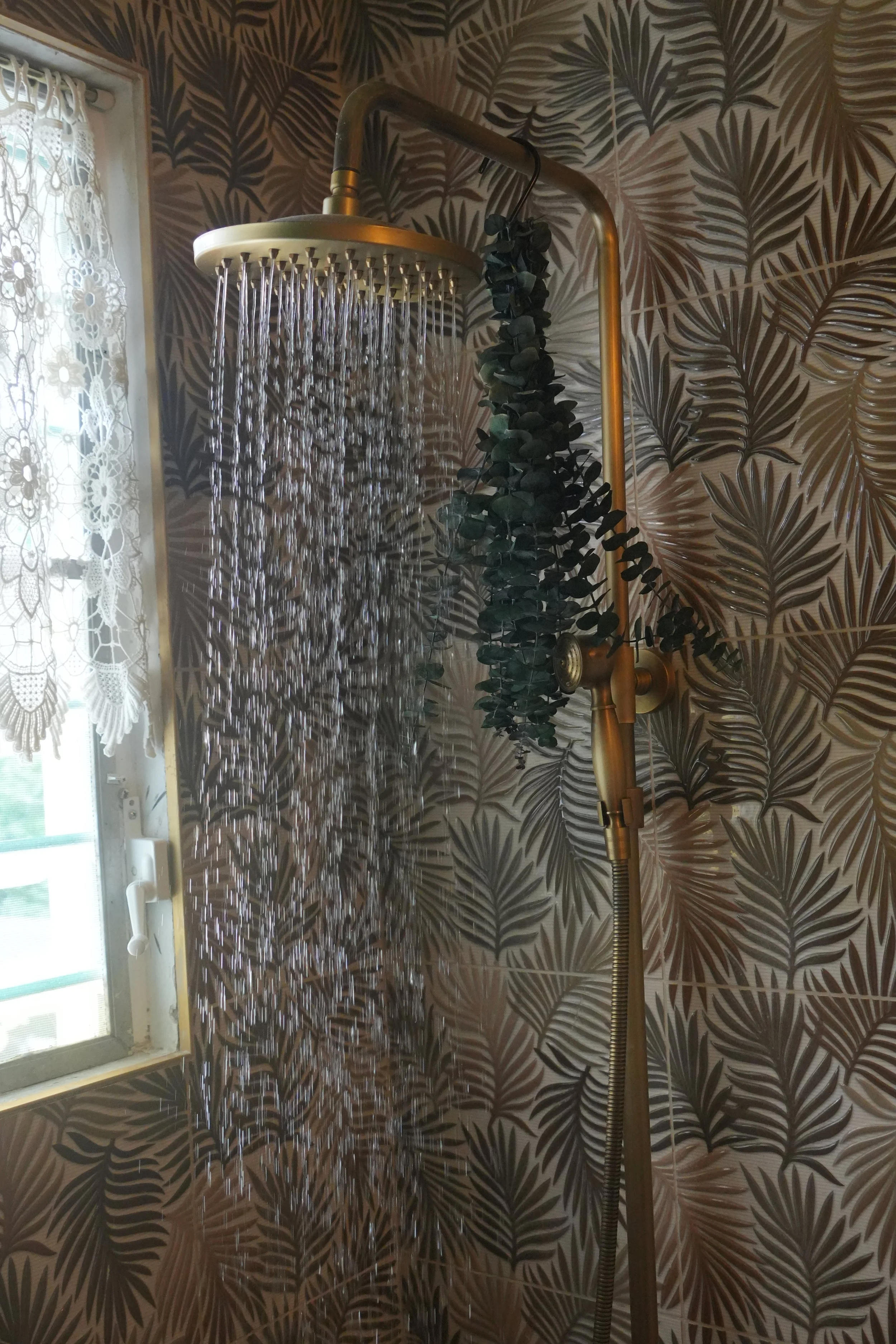 Bathroom shower with rainfall showerhead mounted on a gold-colored metal frame. Water is running from the showerhead. The wall behind is decorated with dark botanical-patterned wallpaper. To the left, there is a window with lace curtains.