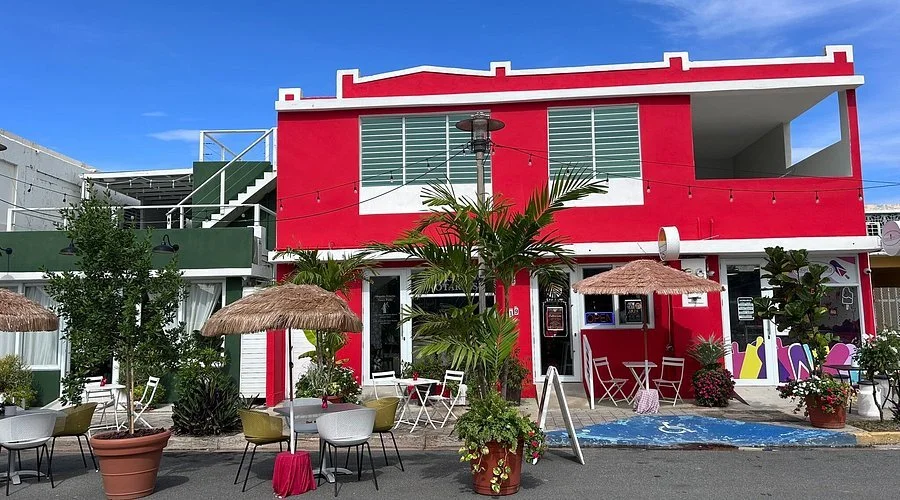 Colorful two-story building painted in red and white, with outdoor seating, tables, plants, and umbrellas, under a bright blue sky.