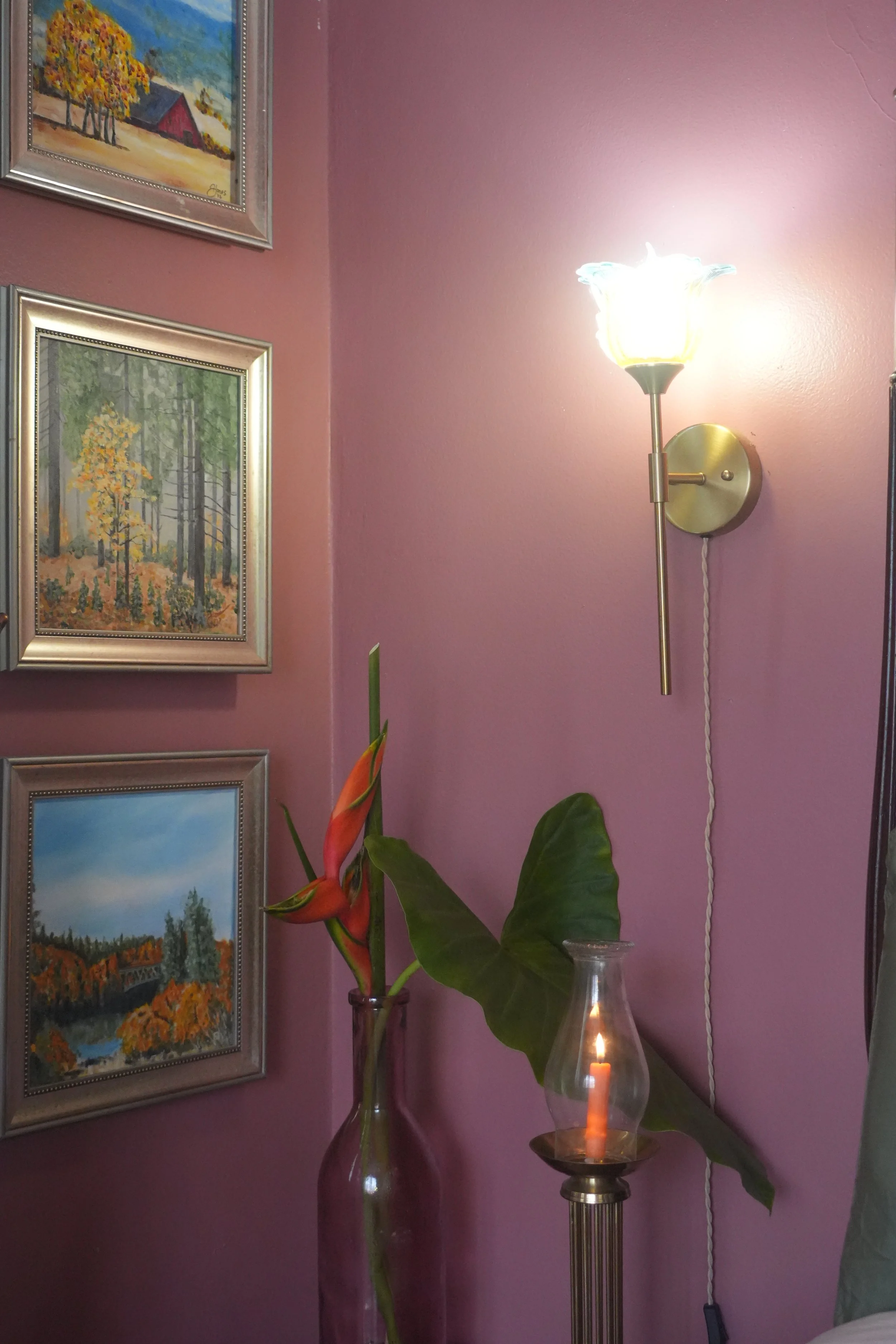 A pink wall decorated with three framed landscape paintings, a tall standing lamp with a flower-shaped light, a glass vase with orange and green flowers, and a candle in a glass holder.