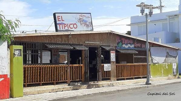 A small wooden restaurant with a sign reading 'El Tipico' on a billboard, located on a sidewalk with a streetlight and palm trees nearby.