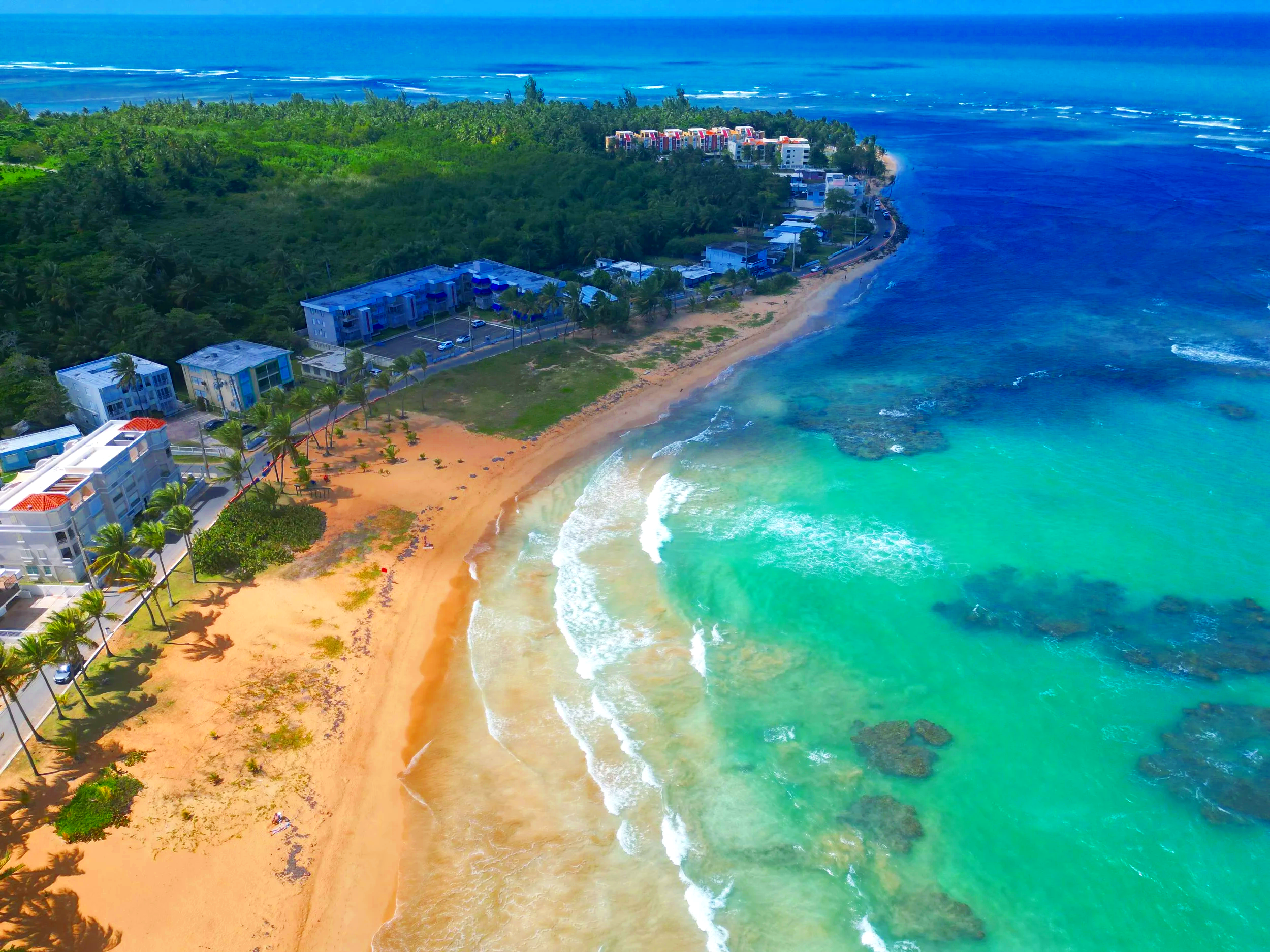 An aerial view of Playa Azul beach with turquoise waters, sandy shoreline, buildings, and a lush green forest in the background.
