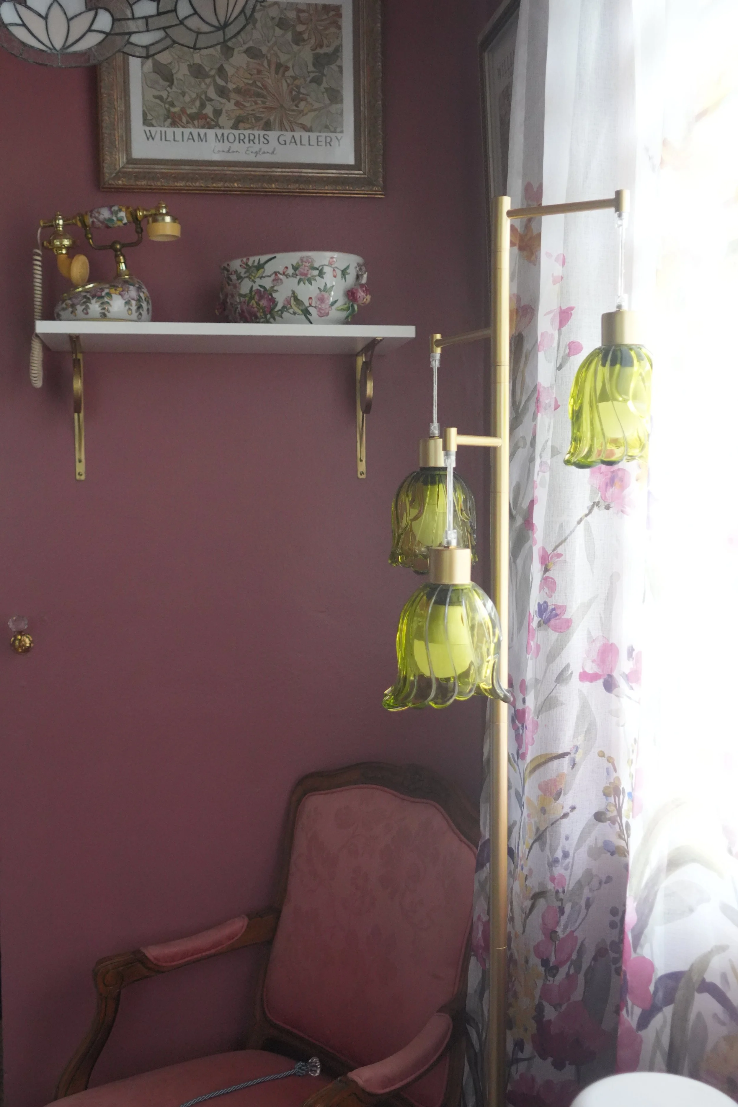 A corner of a room with a pink upholstered armchair, a gold-colored floor lamp with three green glass shades, a curtained window with pink and purple floral patterns, a white shelf with decorative floral pottery, and a framed painting of William Morr