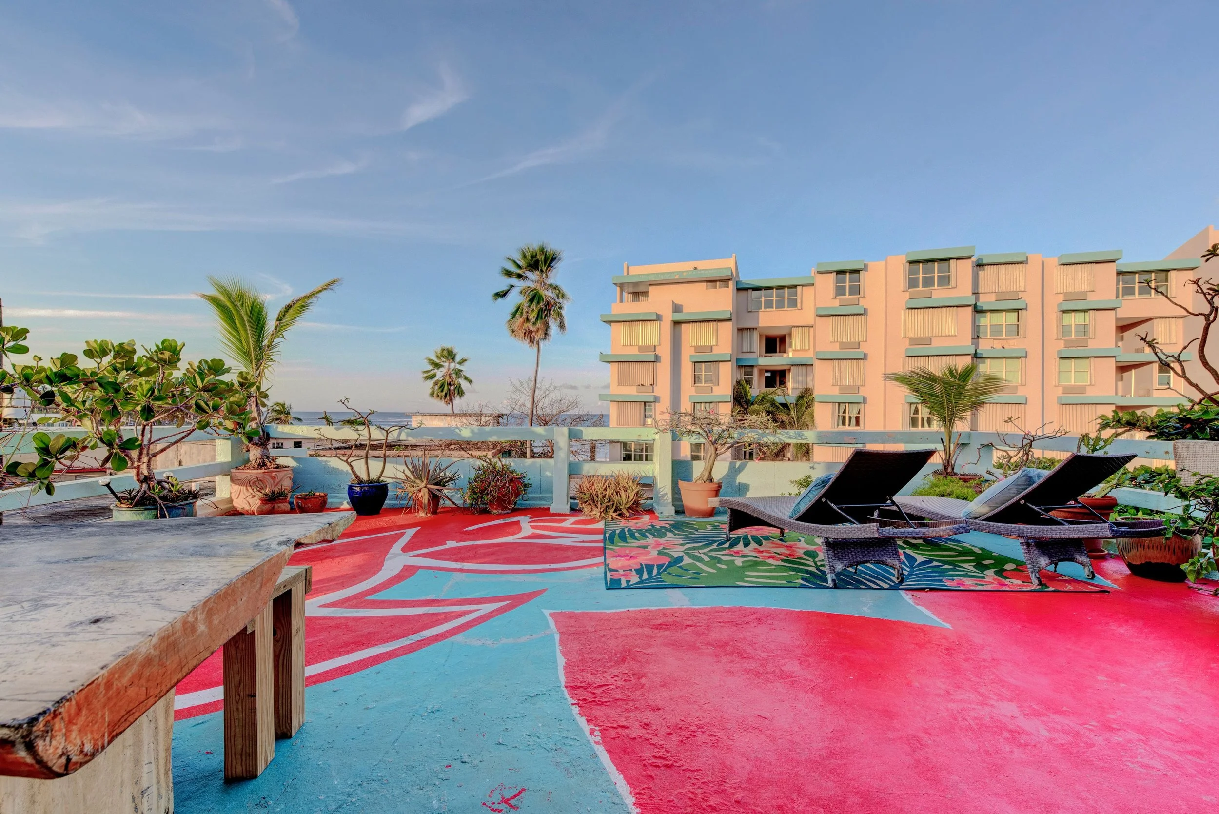 An outdoor rooftop patio with colorful painted flooring featuring pink and blue hues and abstract patterns, decorated with potted plants, two black lounge chairs, and a wooden table, with a pastel-colored building, palm trees, and a partly cloudy blue sky in the background.