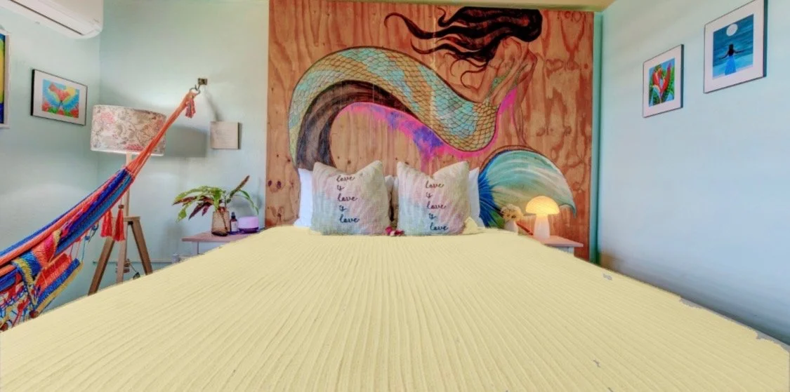 A bedroom with a large bed covered in a light yellow blanket, three pillows with the word 'love' written on them, a colorful mural of a mermaid on the wooden headboard, artwork on the walls, a hammock on the left side, a plant, a lamp, and framed photos.
