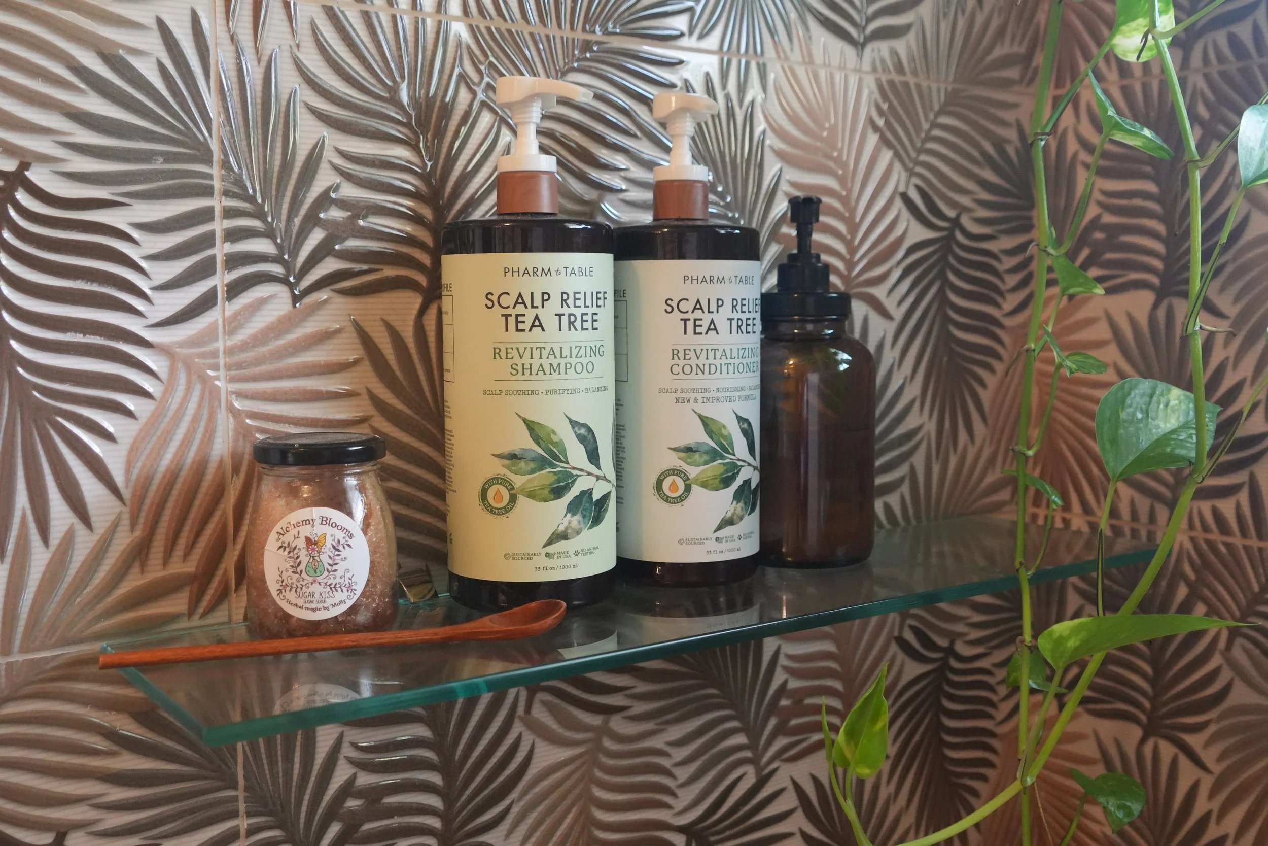 Bathroom shelf with scalp relief tea tree shampoo, scalp relief tea tree conditioner, a jar of Alchemy Blooms Sugar Kiss bath salts, and a small wooden spoon against a leafy-patterned tile background and some green plants.