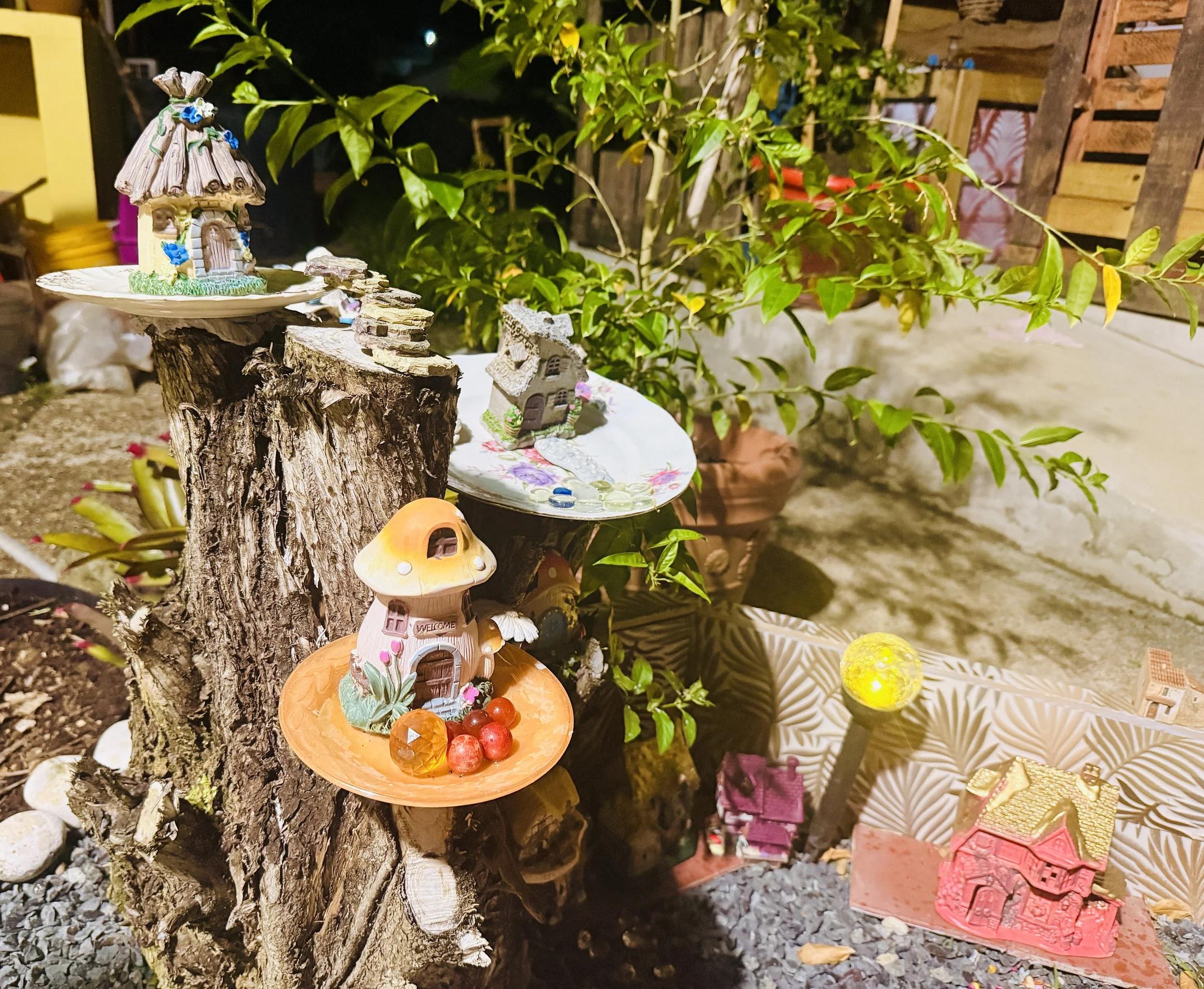 Nighttime outdoor scene with decorative fairy house figurines on a tree stump, surrounded by plants, with a wooden fence and various decorative items in the background.