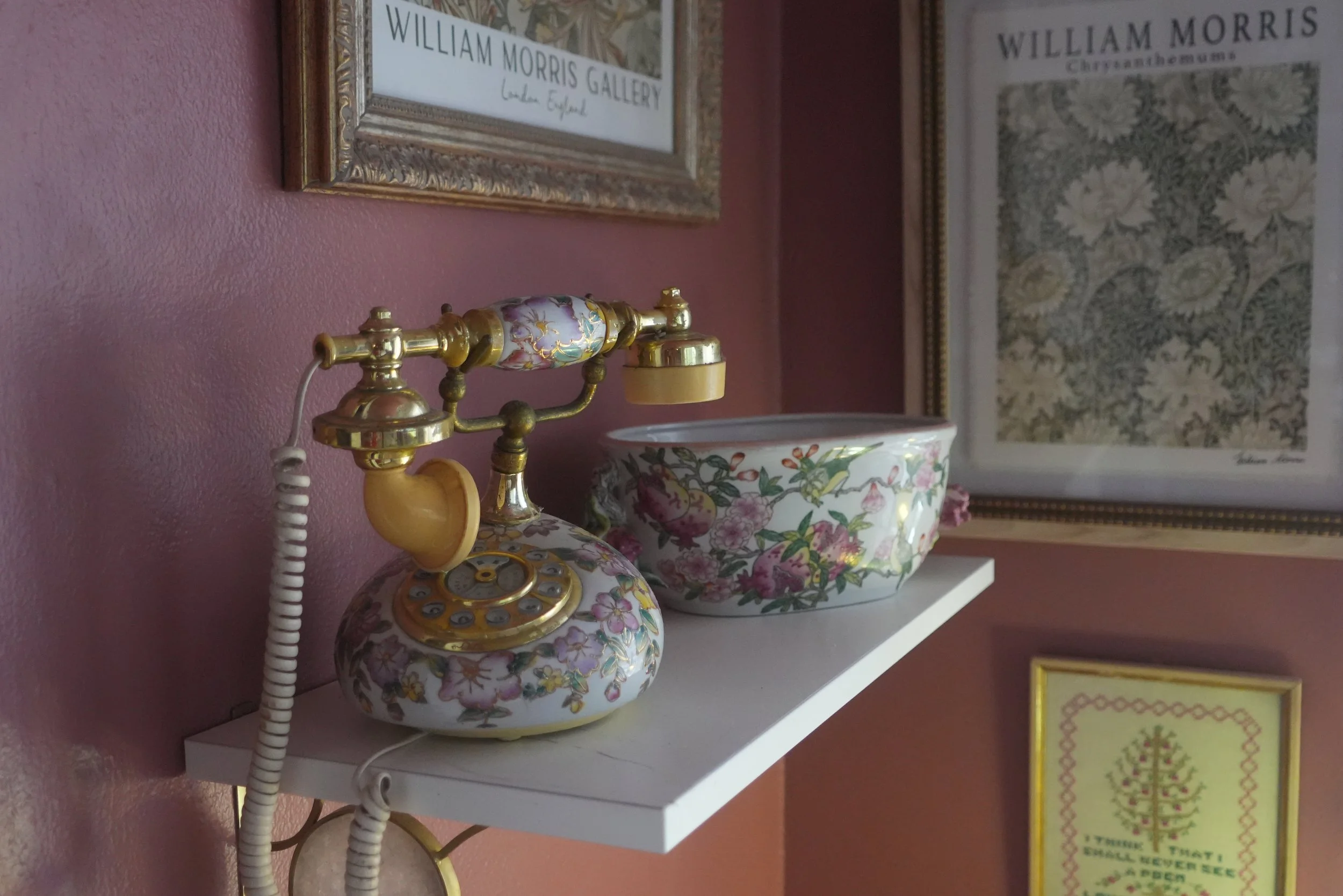Decorative vintage telephone with floral design sitting on a white shelf next to a colorful bowl with a floral pattern, with framed artwork and posters on a pink wall in the background.