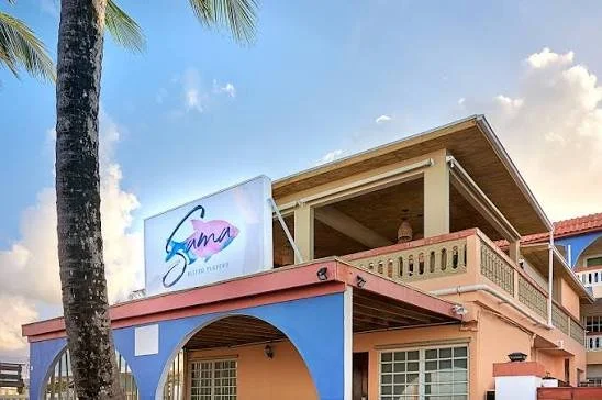 A two-story building with pastel pink and blue exterior, tropical palm trees nearby, and a sign reading 'Samma' on the roof, under a partly cloudy sky.