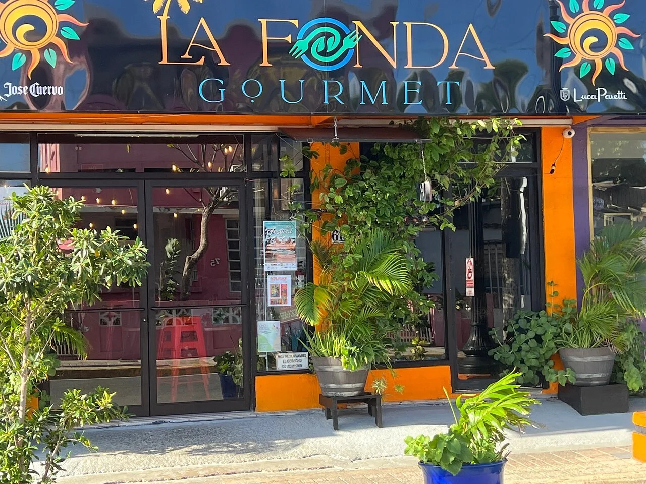 Front of a restaurant named La Fonda Gourmet with plants outside, glass door, and colorful signage.