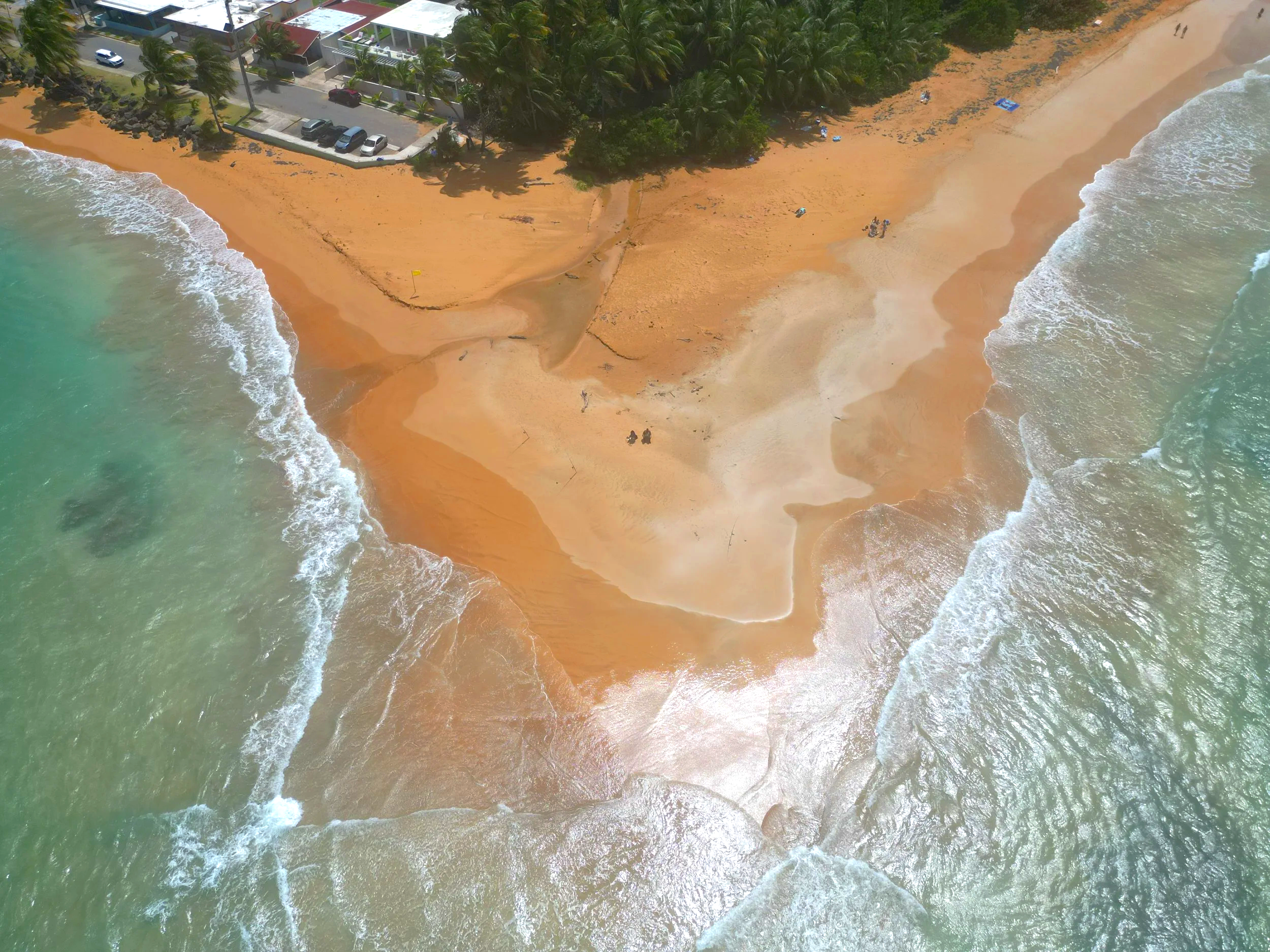Aerial view of the sandy la Punta beach with ocean waves crashing onto the shore, some people and umbrellas on the sand, with a line of trees and parked cars along a street behind the beach.