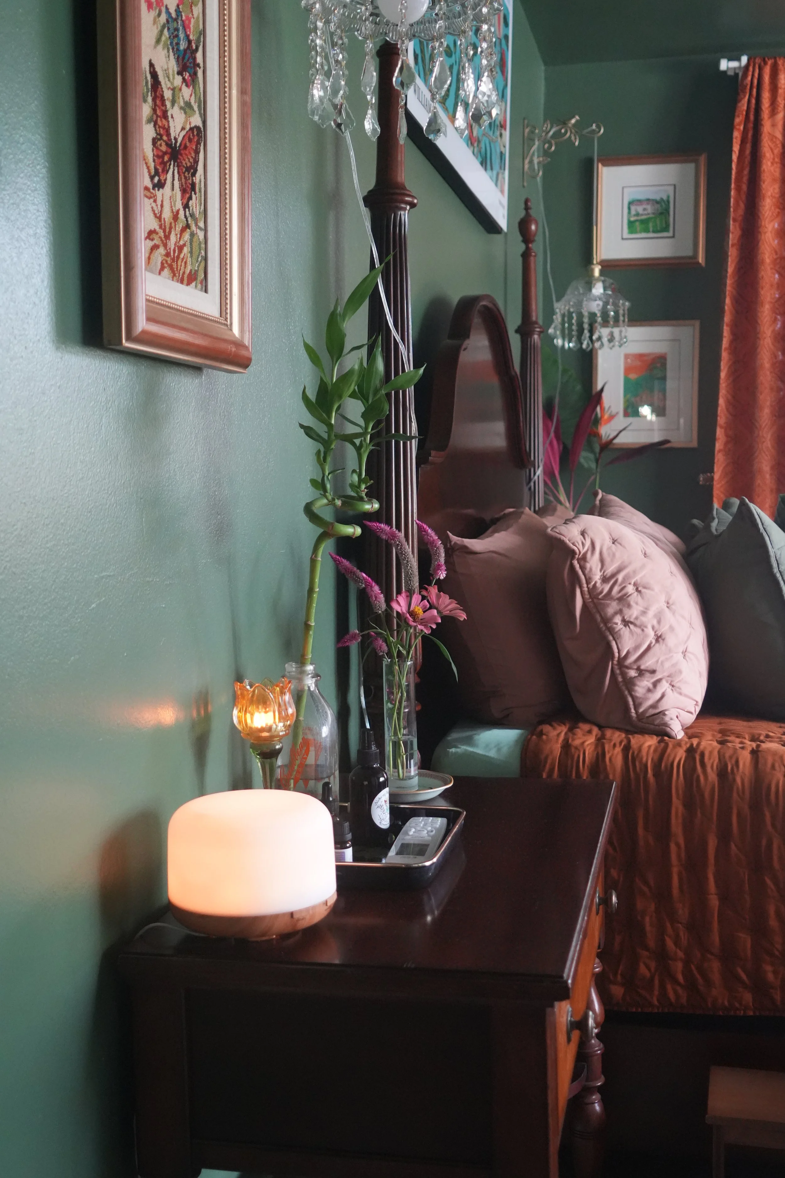 A green wall decorated with framed artwork, a chandelier hanging from the ceiling, and a wooden headboard with pillows. A side table holds a candle and decorative items, and there are various plants on a countertop.