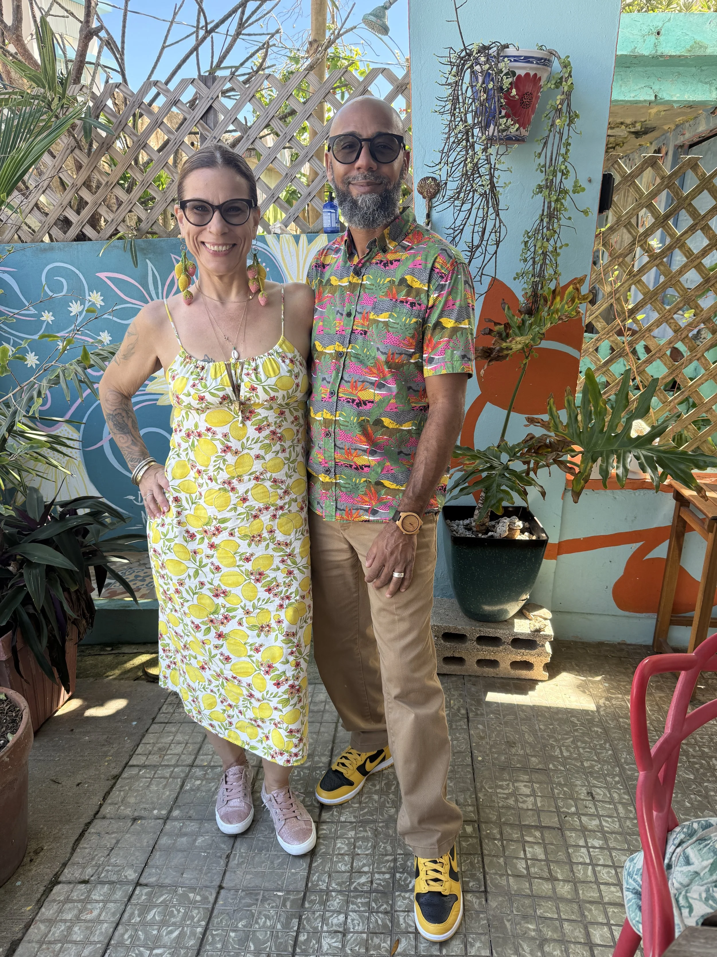 Two people standing in an outdoor patio area. The woman on the left wears a yellow floral dress with lemon prints, pink sneakers, and colorful dangling earrings. The man on the right wears a colorful, patterned short-sleeve shirt, beige pants, and yellow and black sneakers. They are smiling, and behind them are decorative potted plants, a painted gallery wall, and a lattice fence.