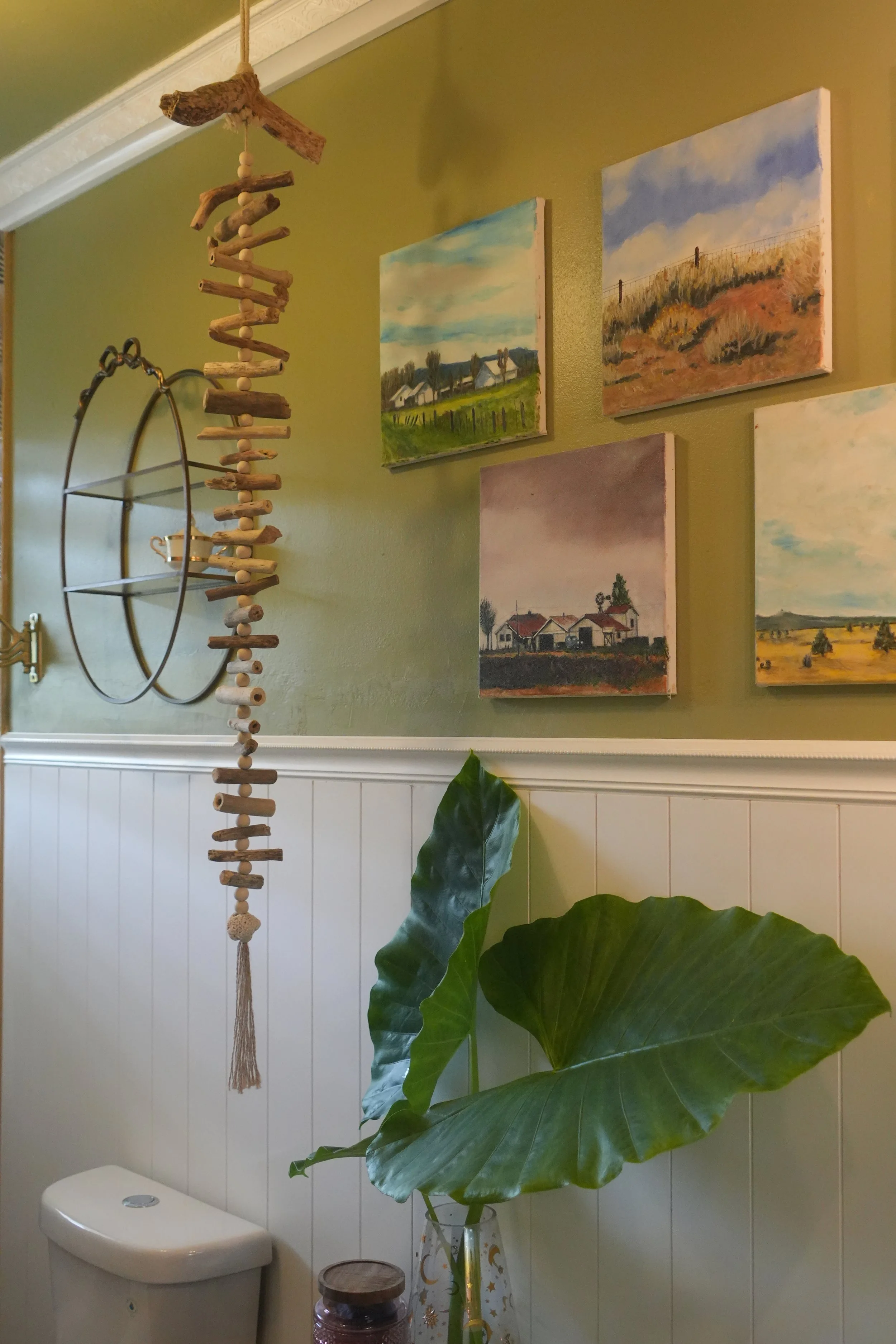 A green wall with four landscape paintings, a large leafy houseplant in a decorative vase, and a hanging wall decor made of driftwood and wooden beads in a room, possibly a bathroom.