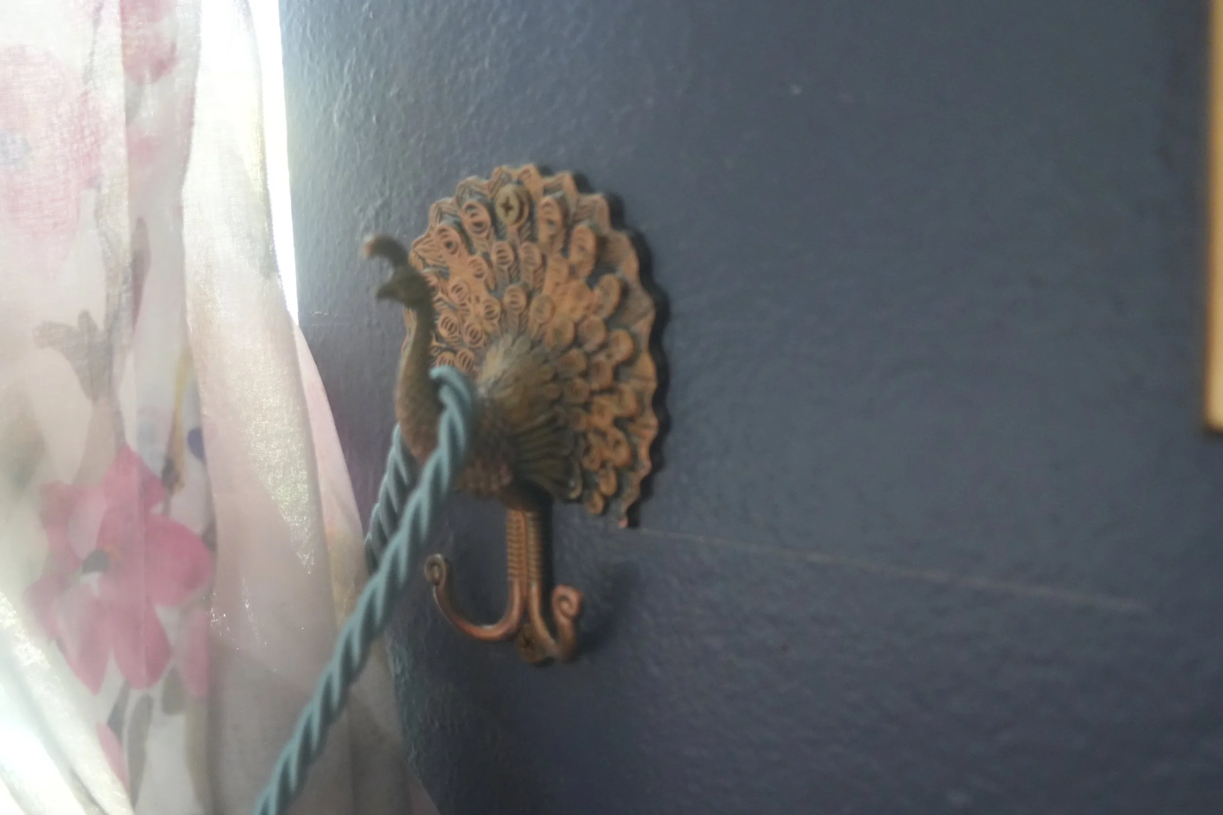 Decorative metal decorative piece on a dark blue wall with a twisted cord hanging from it, next to a curtain with a floral pattern.