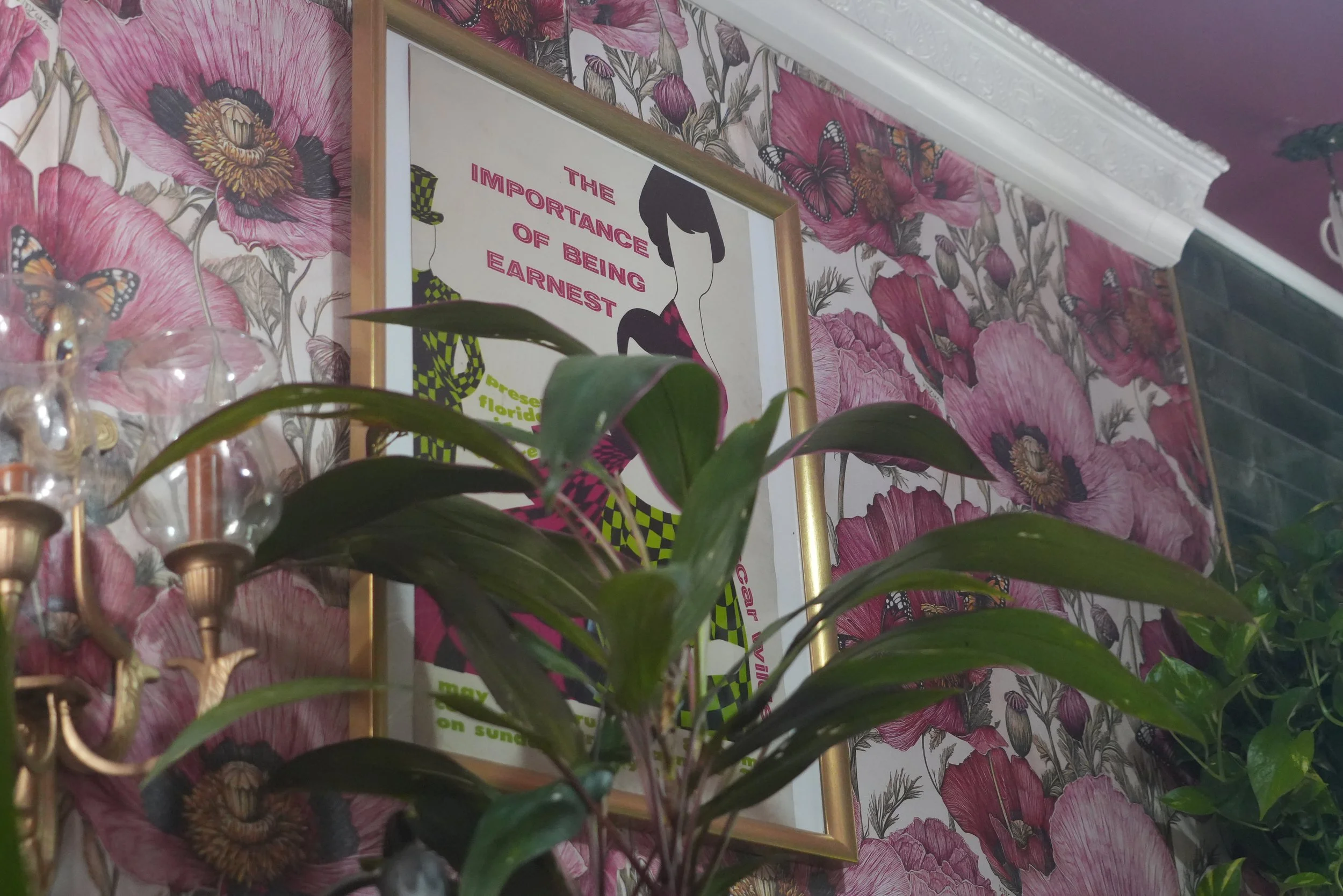 Interior wall covered with floral wallpaper, displaying a framed poster that reads 'The importance of being earnest' with a stylized woman and snake illustration, flower-shaped light fixtures in the foreground.