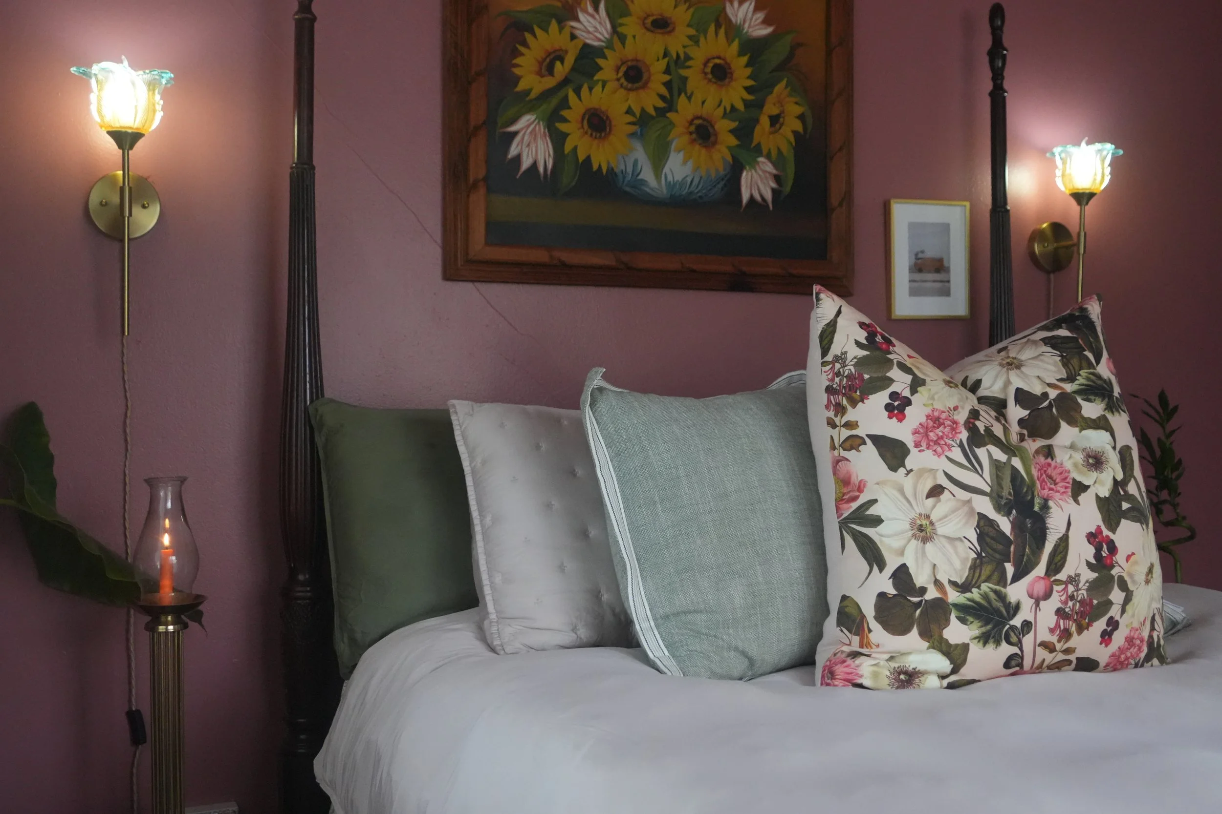 A bedroom with a dark metal four-poster bed, a large rectangular painting of sunflowers on a wooden frame hanging on a pink wall, and two brass wall sconces with floral-shaped glass shades on either side of the painting. The bed has a white bedspread