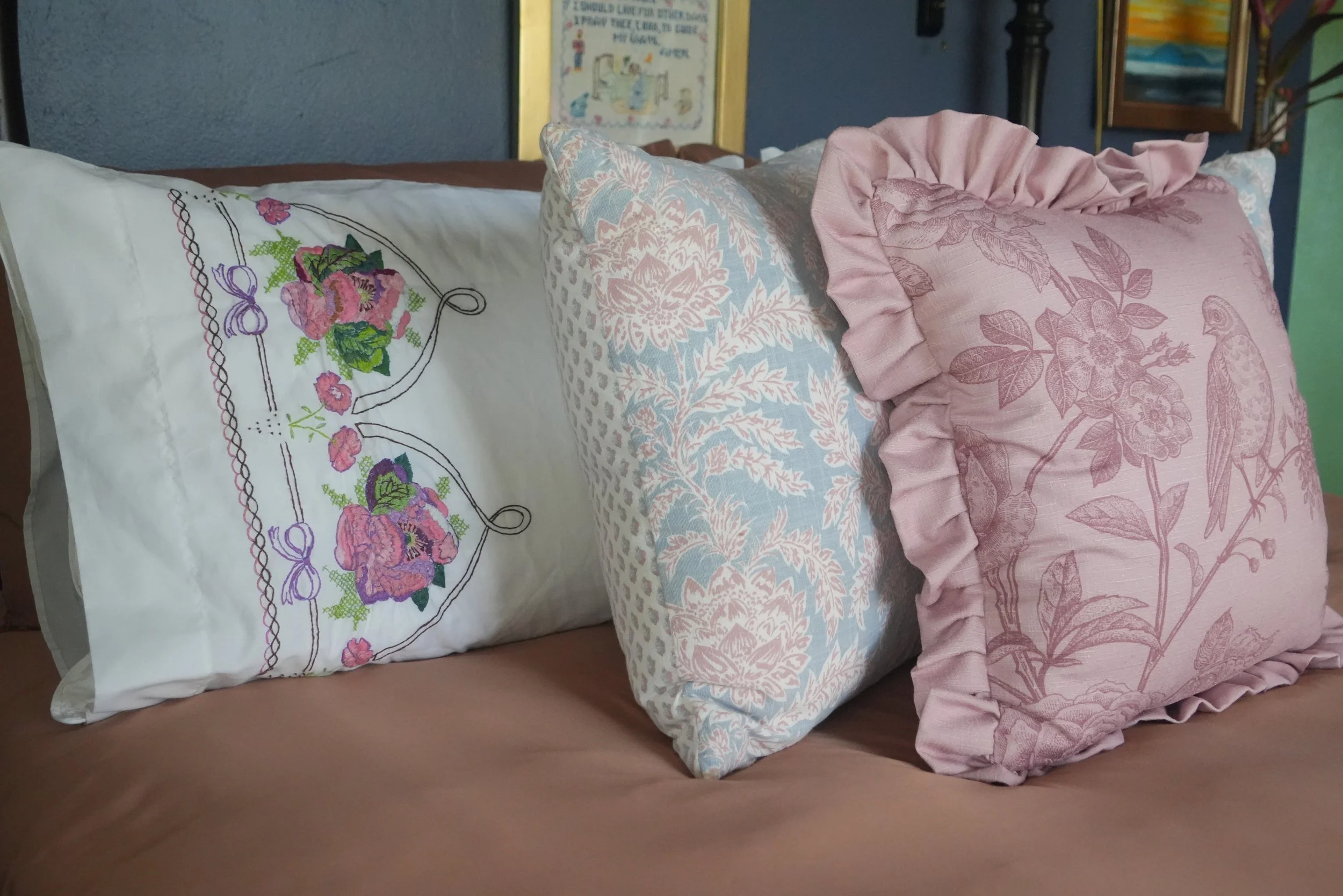 A bed with multiple decorative pillows, including a large white pillow with embroidered flowers, a light blue pillow with a floral pattern, and a pink pillow with a ruffled edge and floral design. The background shows framed artwork on the wall.